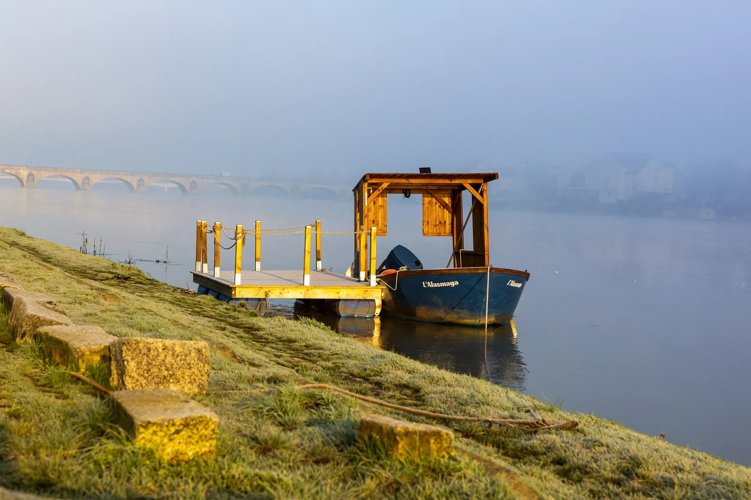 Blue river boat moored at a yellow floating dock, frost on the grass bank, arched stone bridge fading into morning mist behind, Saumur