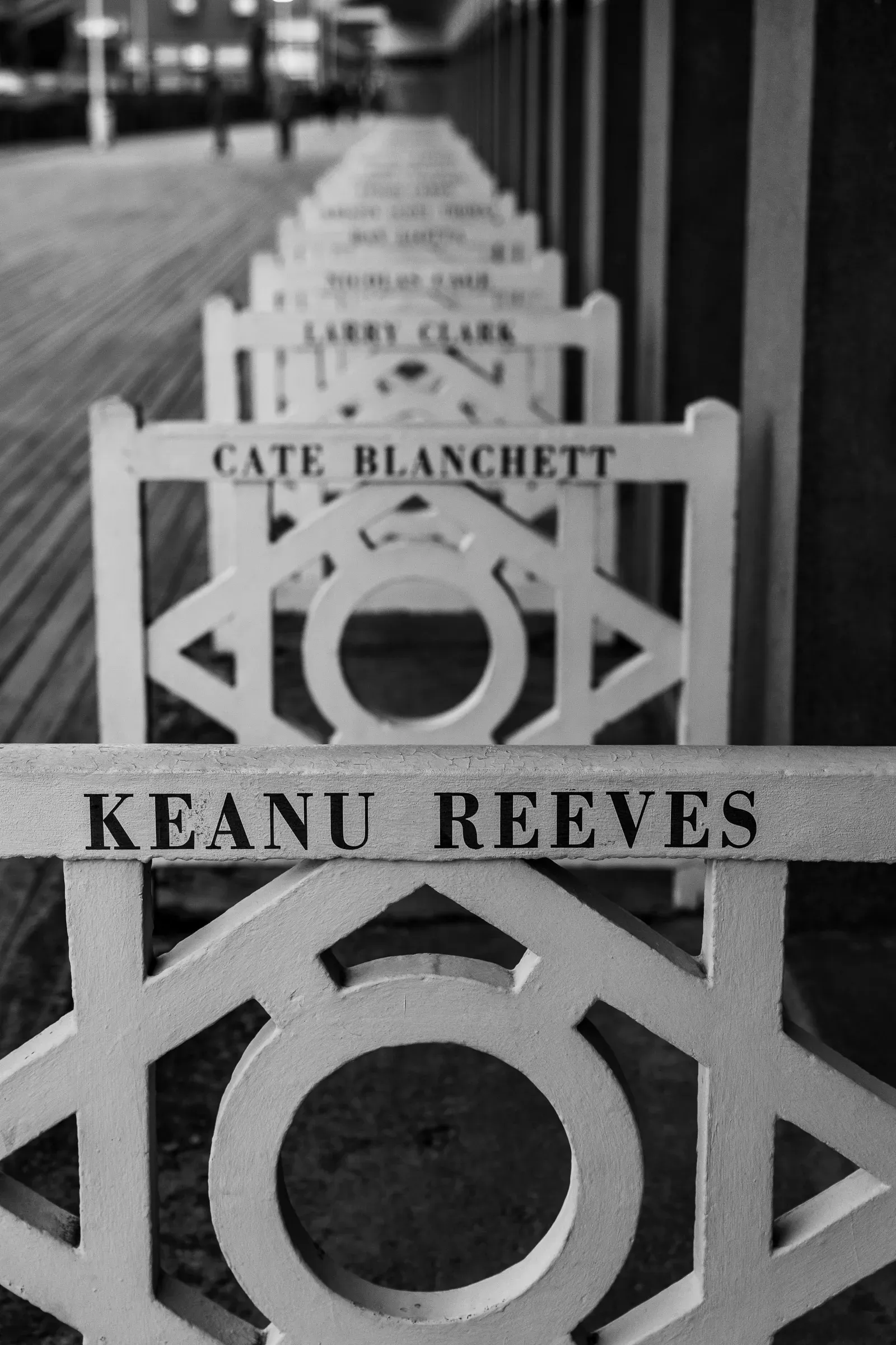 Black and white — row of wooden boardwalk chairs stenciled with celebrity names, Keanu Reeves in the foreground, Cate Blanchett and Larry Clark behind, receding into the distance, Trouville