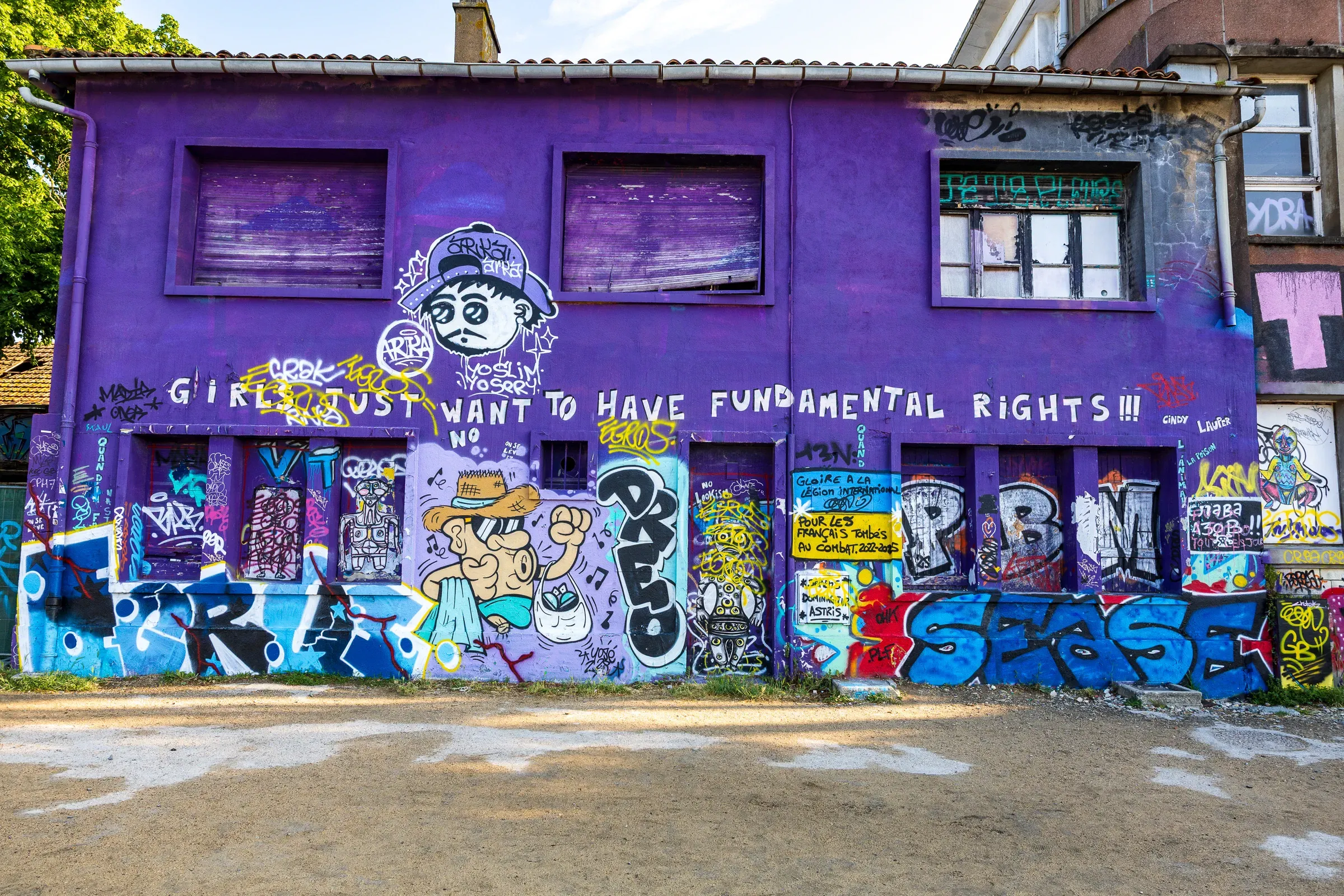 Purple building covered in graffiti, large white text reading 'Girls just want to have fundamental rights!!!' above cartoon figures and colorful tags in blue, red, and yellow