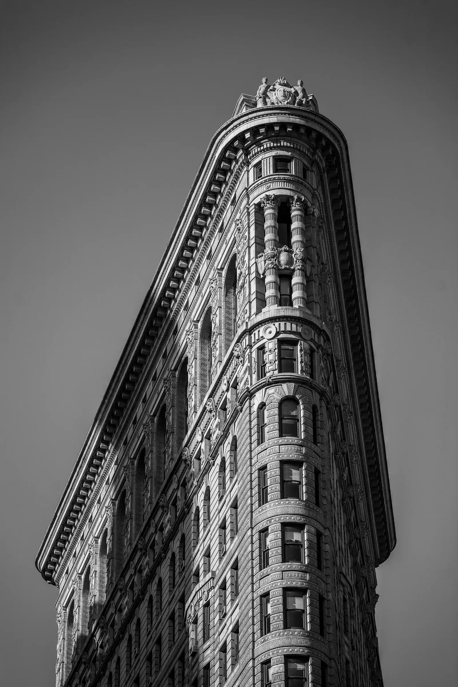 The Flatiron Building in New York shot from street level looking up, the narrow limestone prow tapering to a decorative crown against a pale grey sky