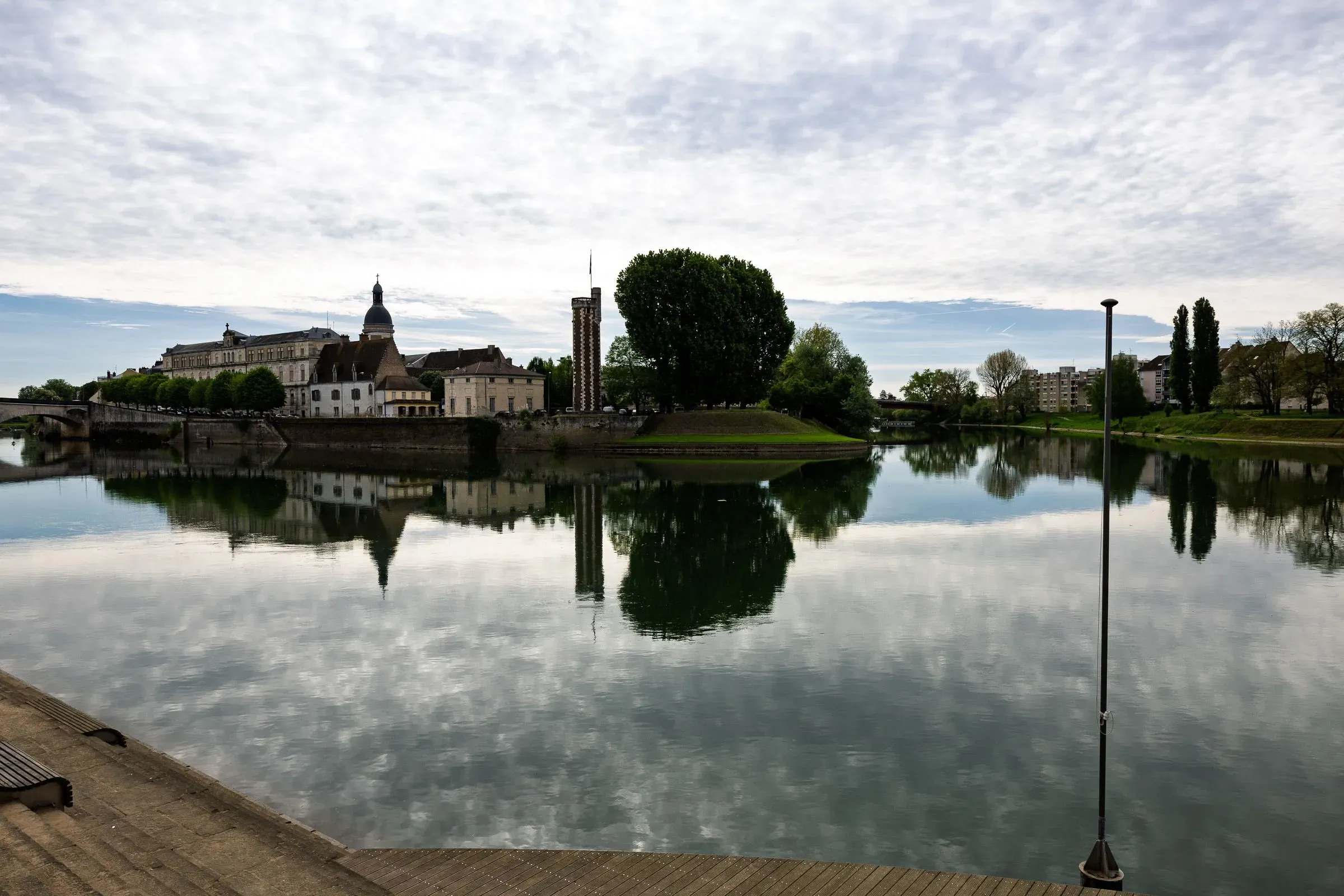 The Saône river with a domed building and bell tower on the quay, a bridge and lamppost in the foreground, overcast sky, Chalon-sur-Saône