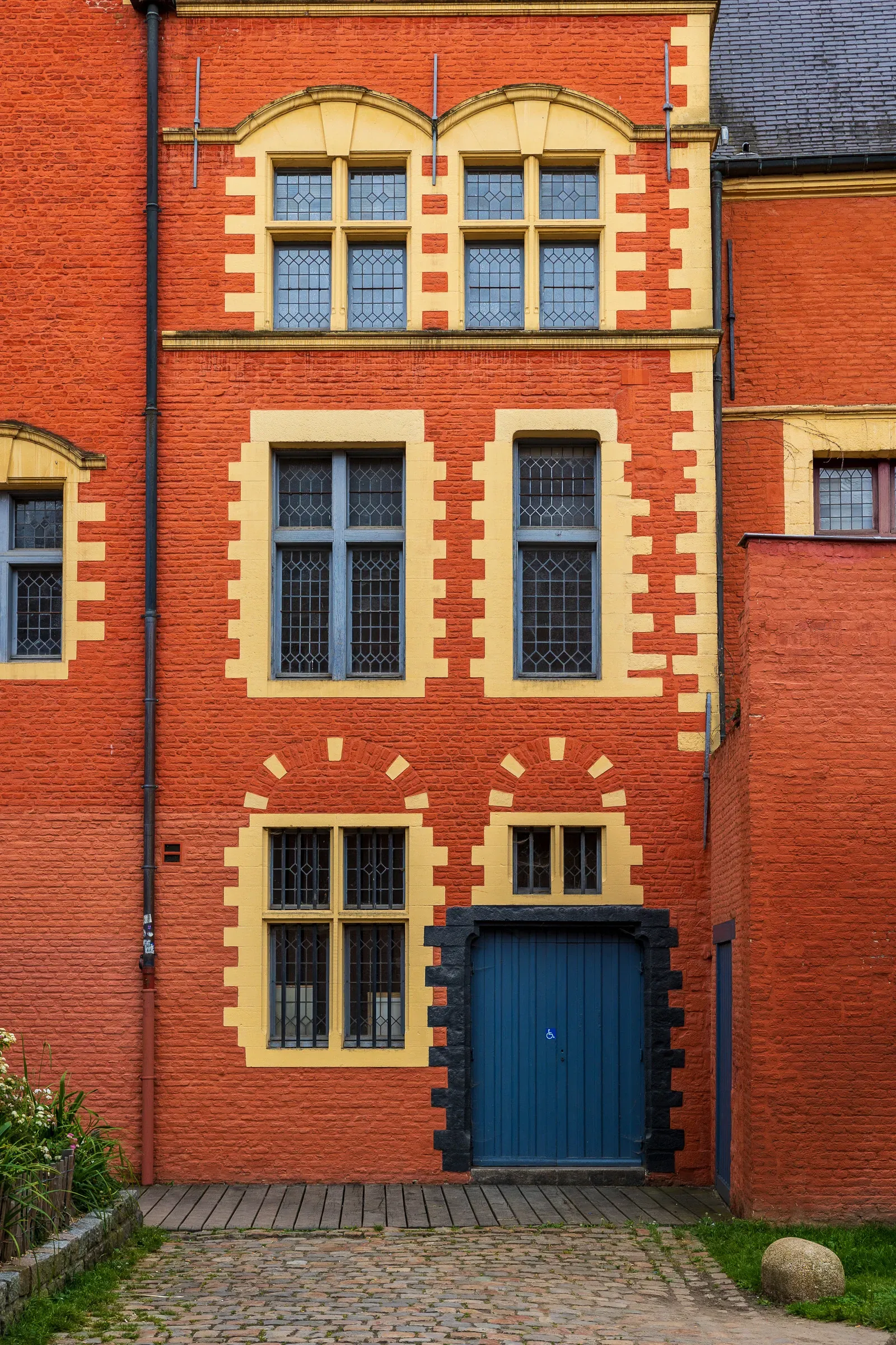 Red brick facade with yellow stone arched windows and a blue door set on cobblestones, Lille