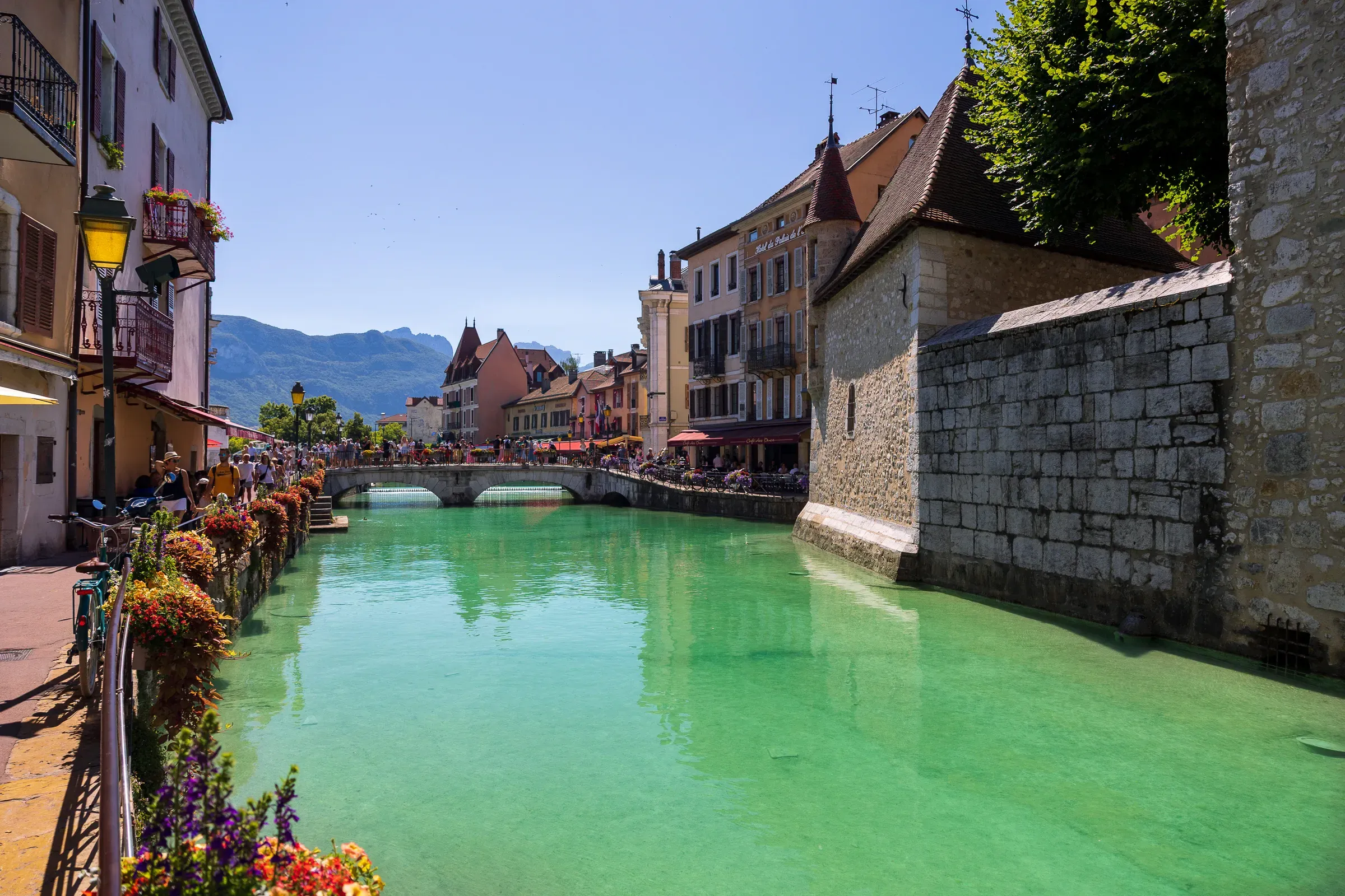 The Thiou canal lined with colorful buildings and flower-covered railings, alpine mountains visible beyond, Annecy