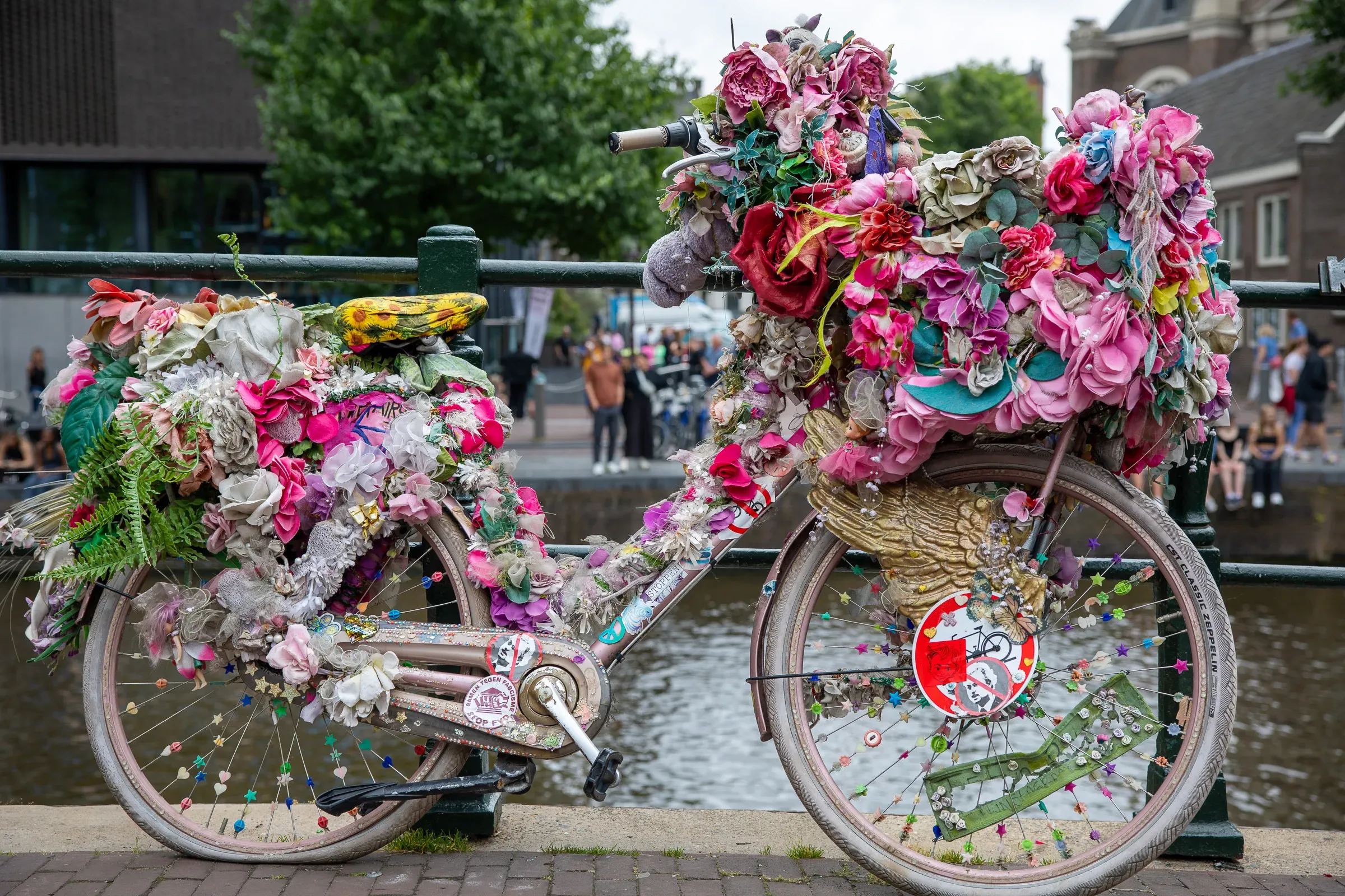 A bike completely covered in pink, red, and mixed fabric flowers, chained to a bridge railing above an Amsterdam canal