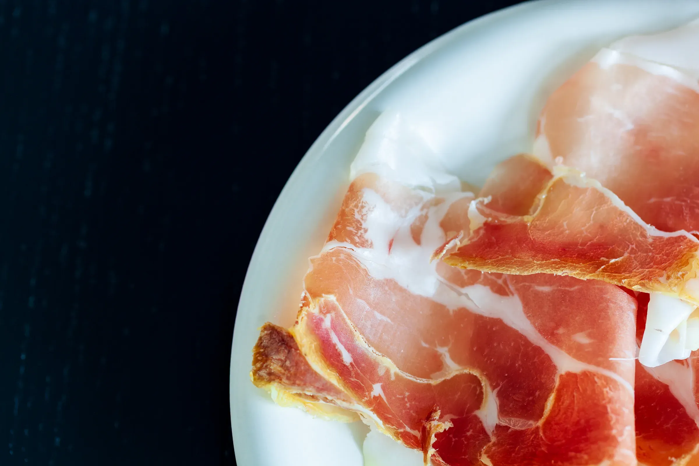 Thin slices of cured ham on a white plate, fat marbled through the meat, dark background — partial overhead close-up