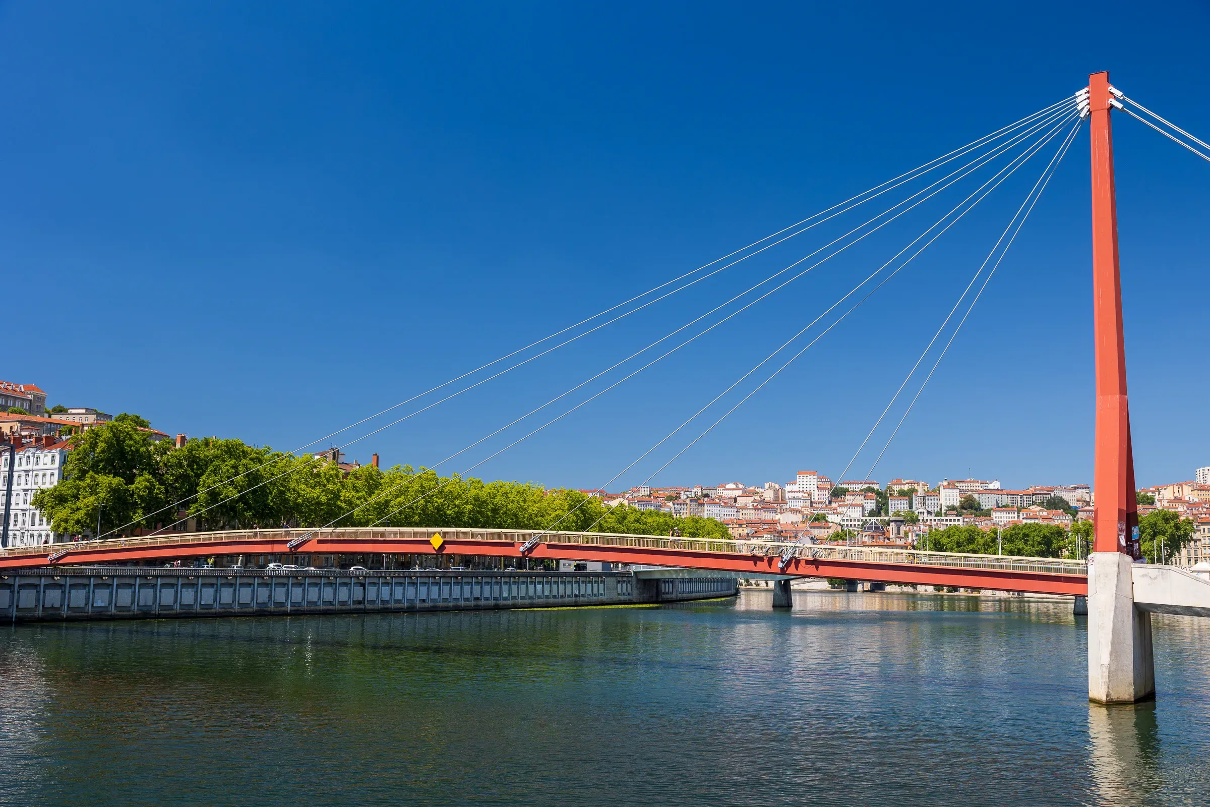 Red cable-stayed pedestrian bridge over the Saône river under a clear blue sky, Lyon