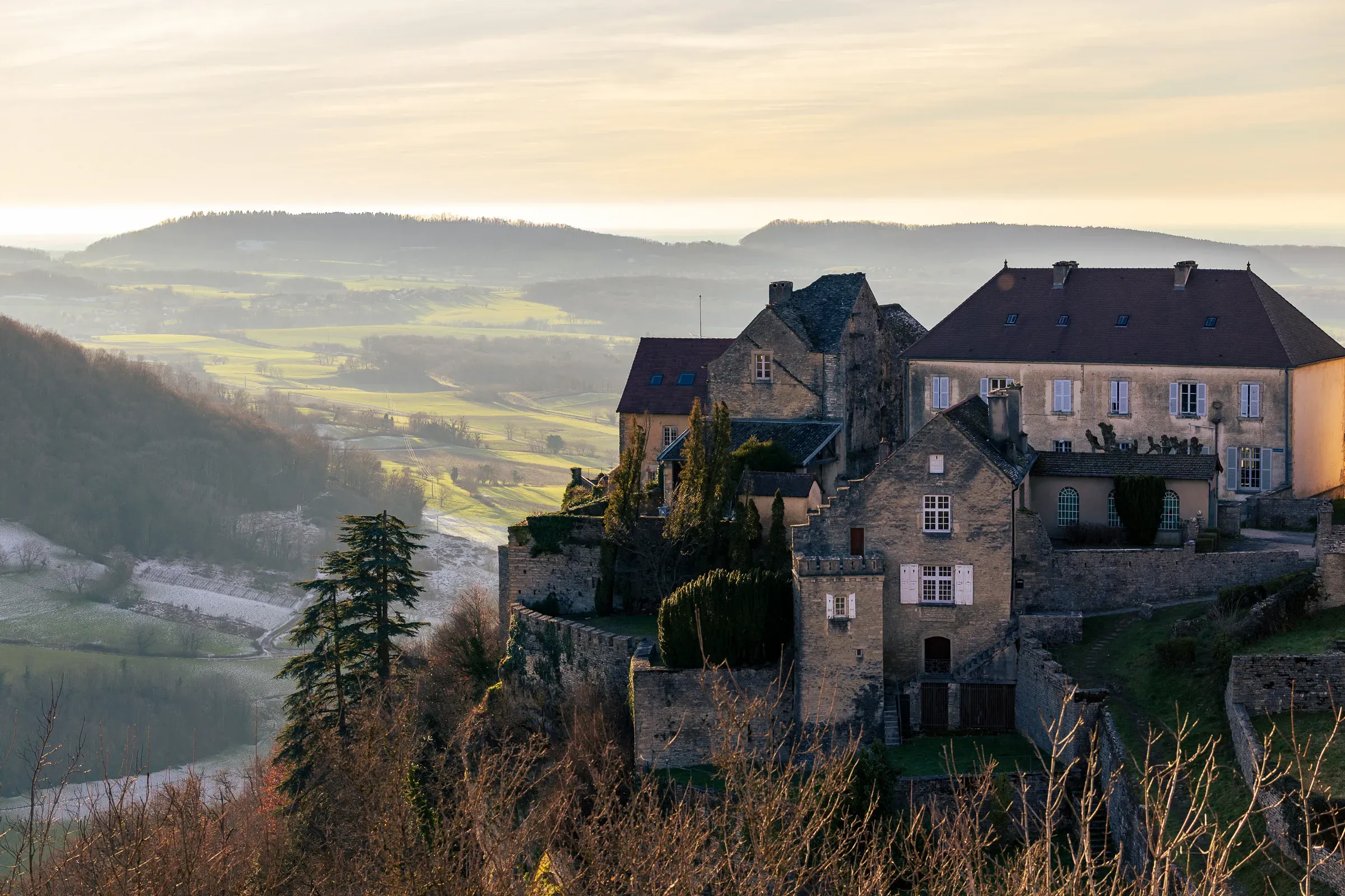 Hilltop medieval château overlooking a misty Jura valley in soft golden morning light, bare trees in the foreground