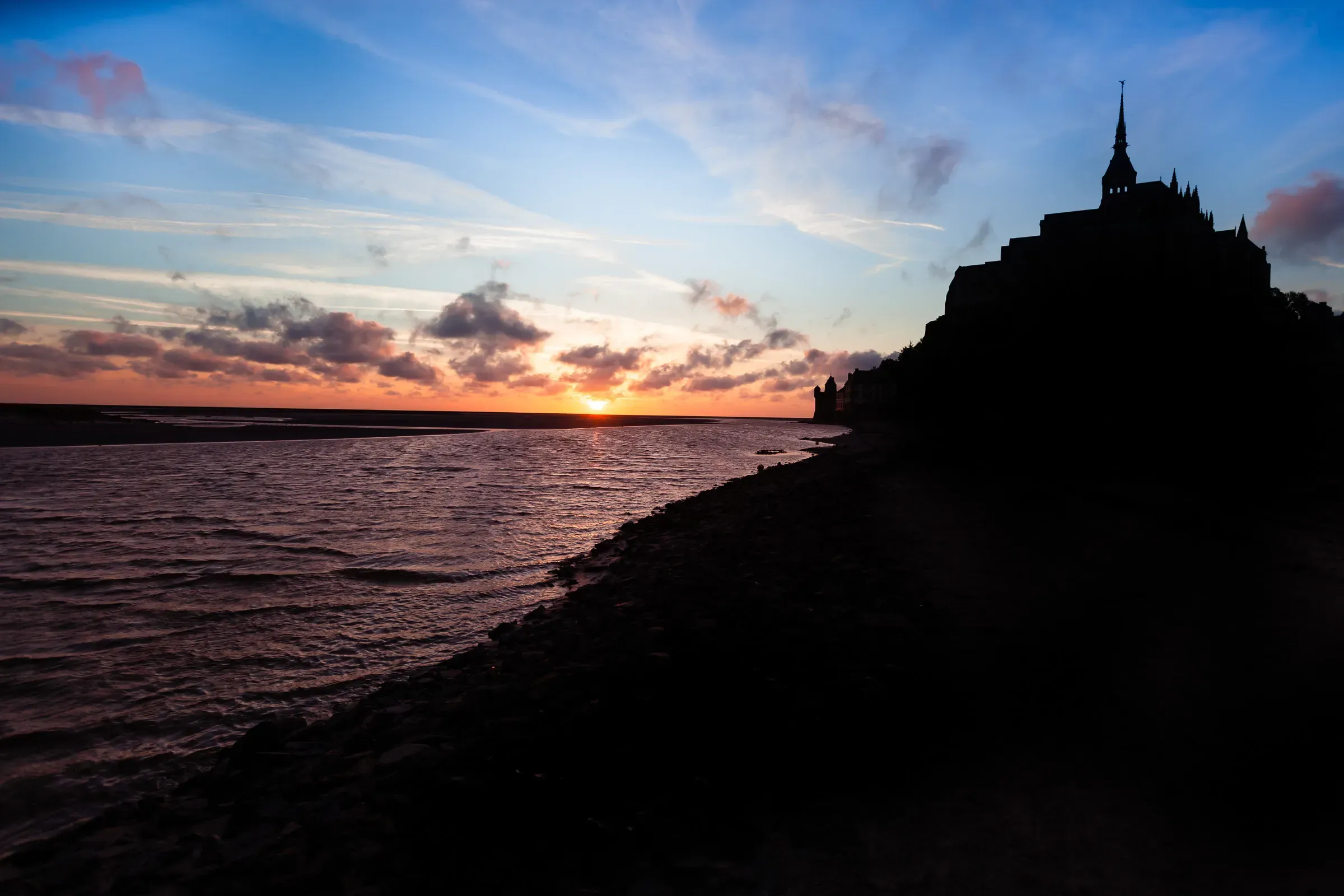 Mont Saint-Michel silhouetted against a sunset sky — orange horizon, pink clouds, dark water of the bay in the foreground