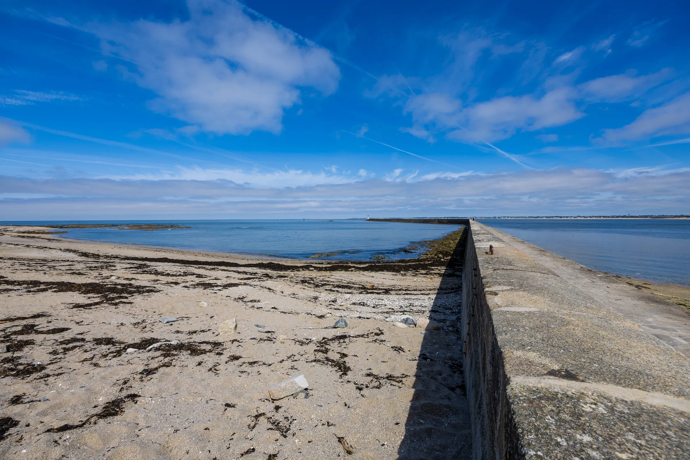 Long stone seawall stretching to the horizon under a bright blue sky, sandy beach with seaweed on the left