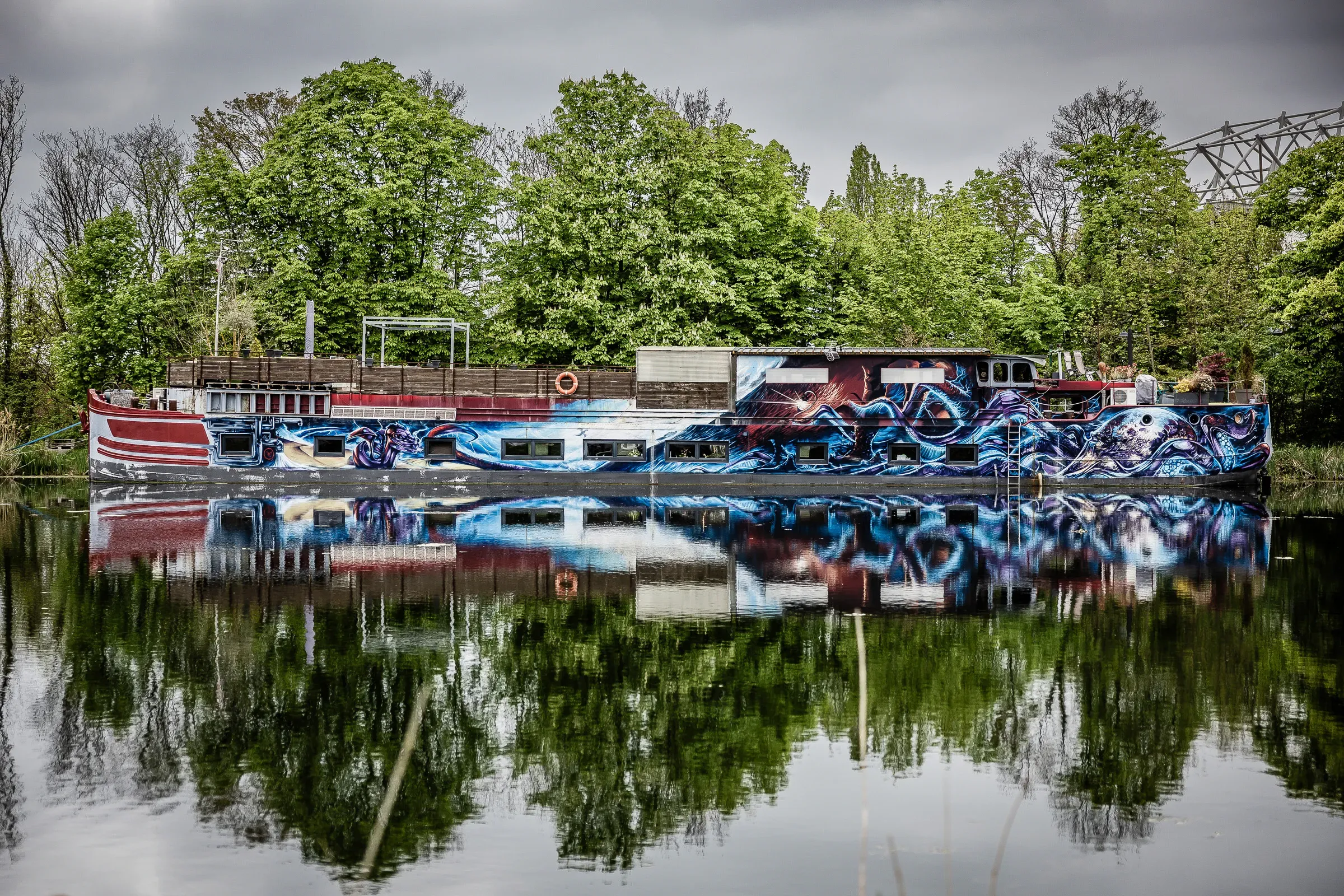 A barge entirely covered in a blue, red, and white graffiti mural, moored on the Moselle river, perfectly mirrored in still water