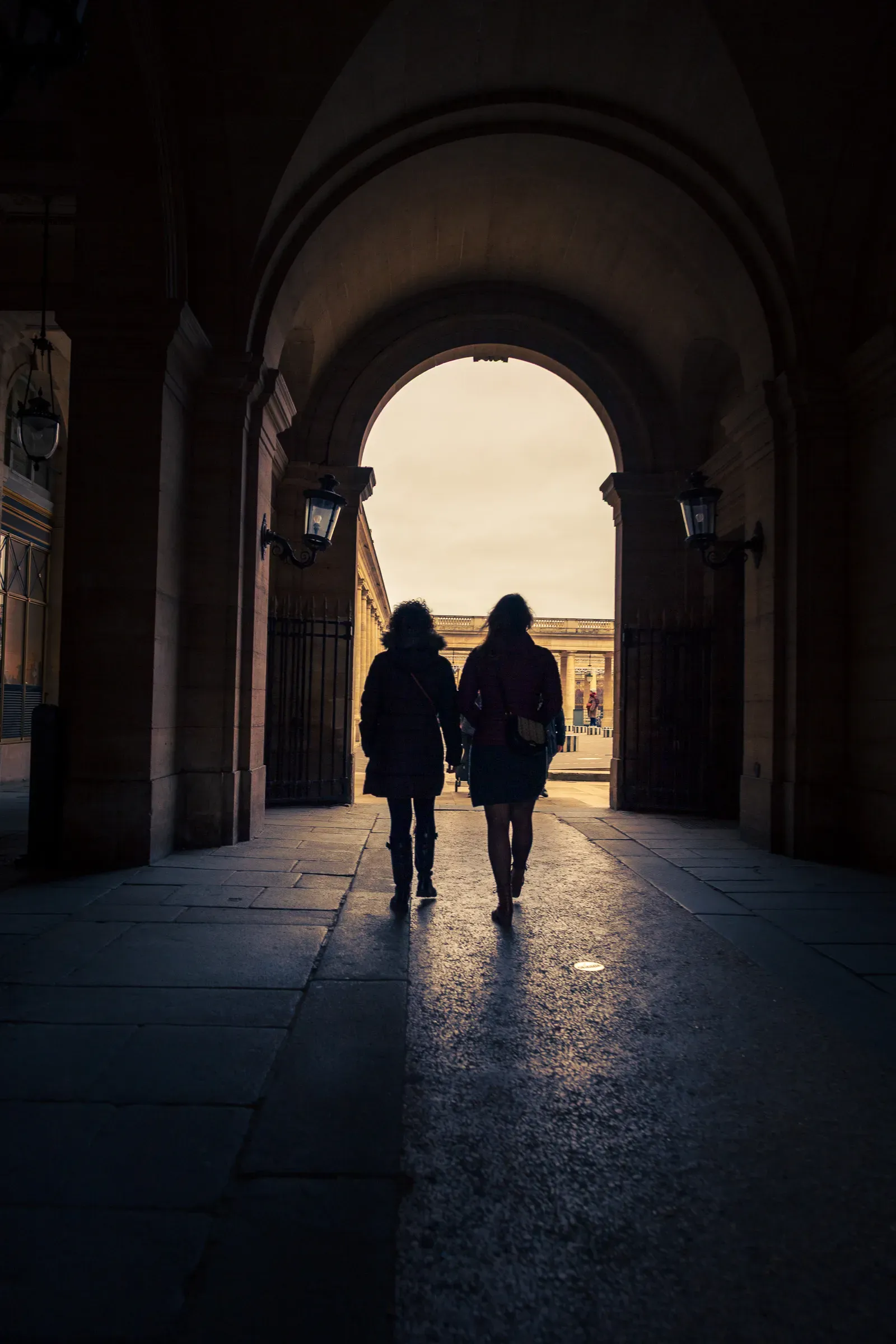 Two people walking away through a stone archway, backlit by daylight, silhouetted against a colonnaded courtyard beyond