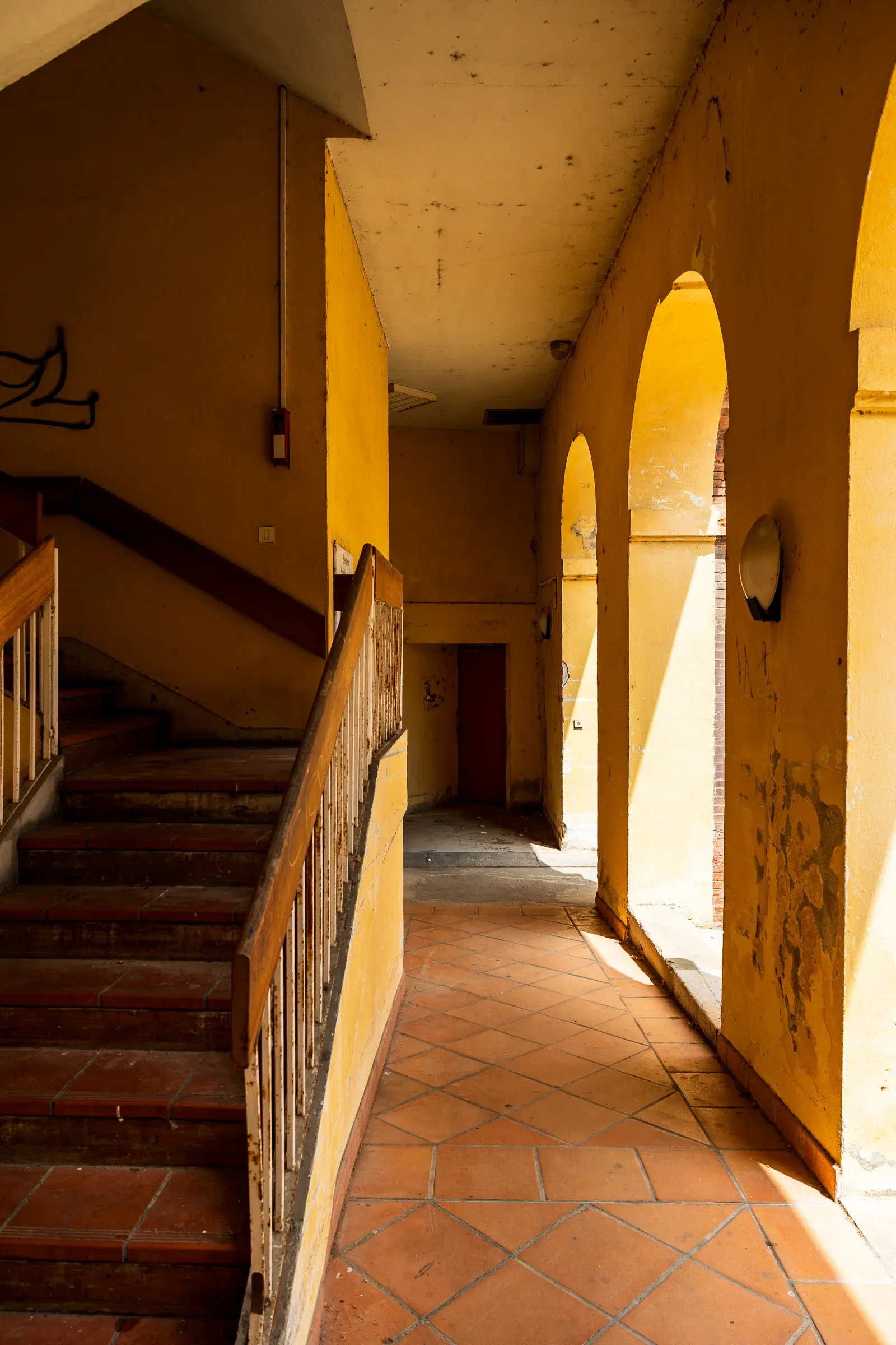 Sunlit interior arcade with ochre-yellow walls, arched openings casting sharp light onto a terracotta tile floor, wooden staircase rising on the left, Toulouse
