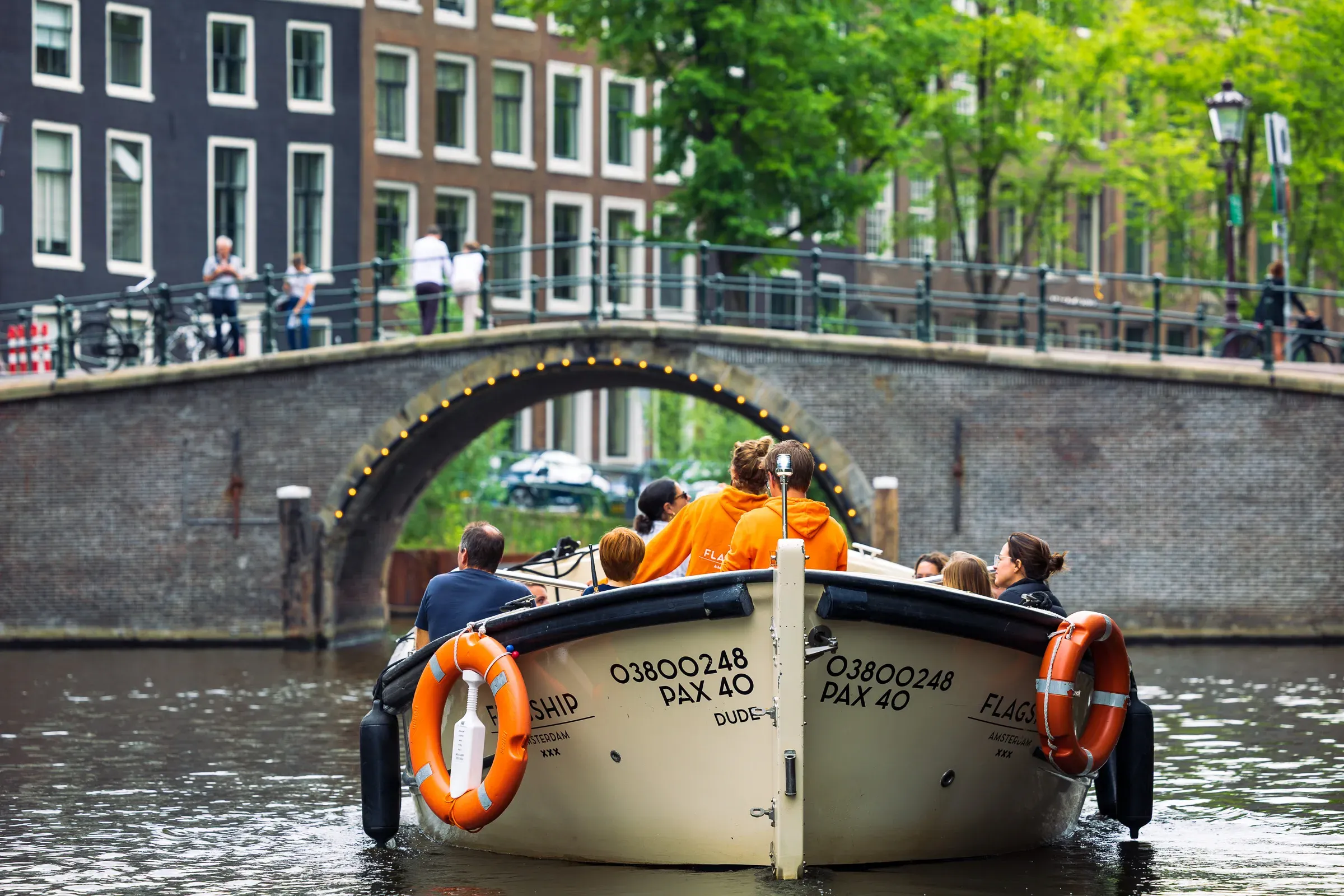 Canal tour boat with passengers in orange jackets heading through a lit brick arch bridge, Amsterdam