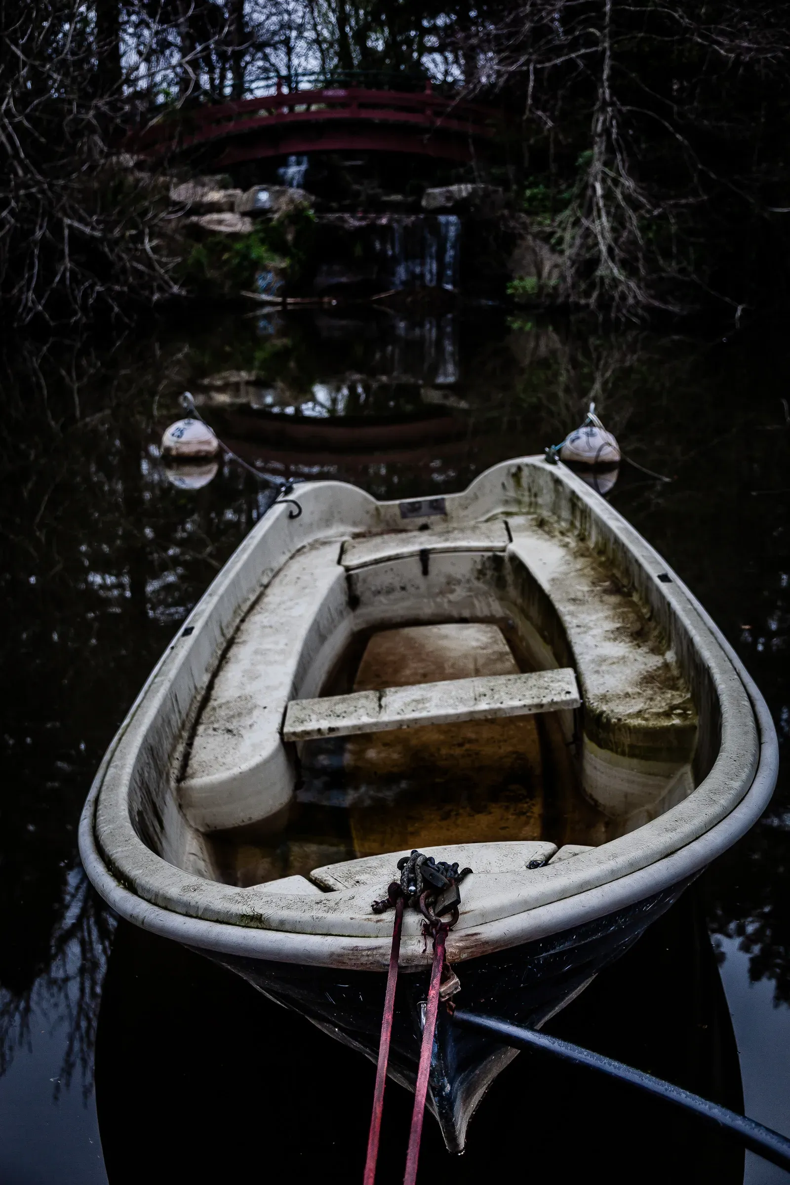 Interior of an old deteriorated rowing boat seen from the bow, weathered bench seats, a red arched bridge reflected in the dark water behind