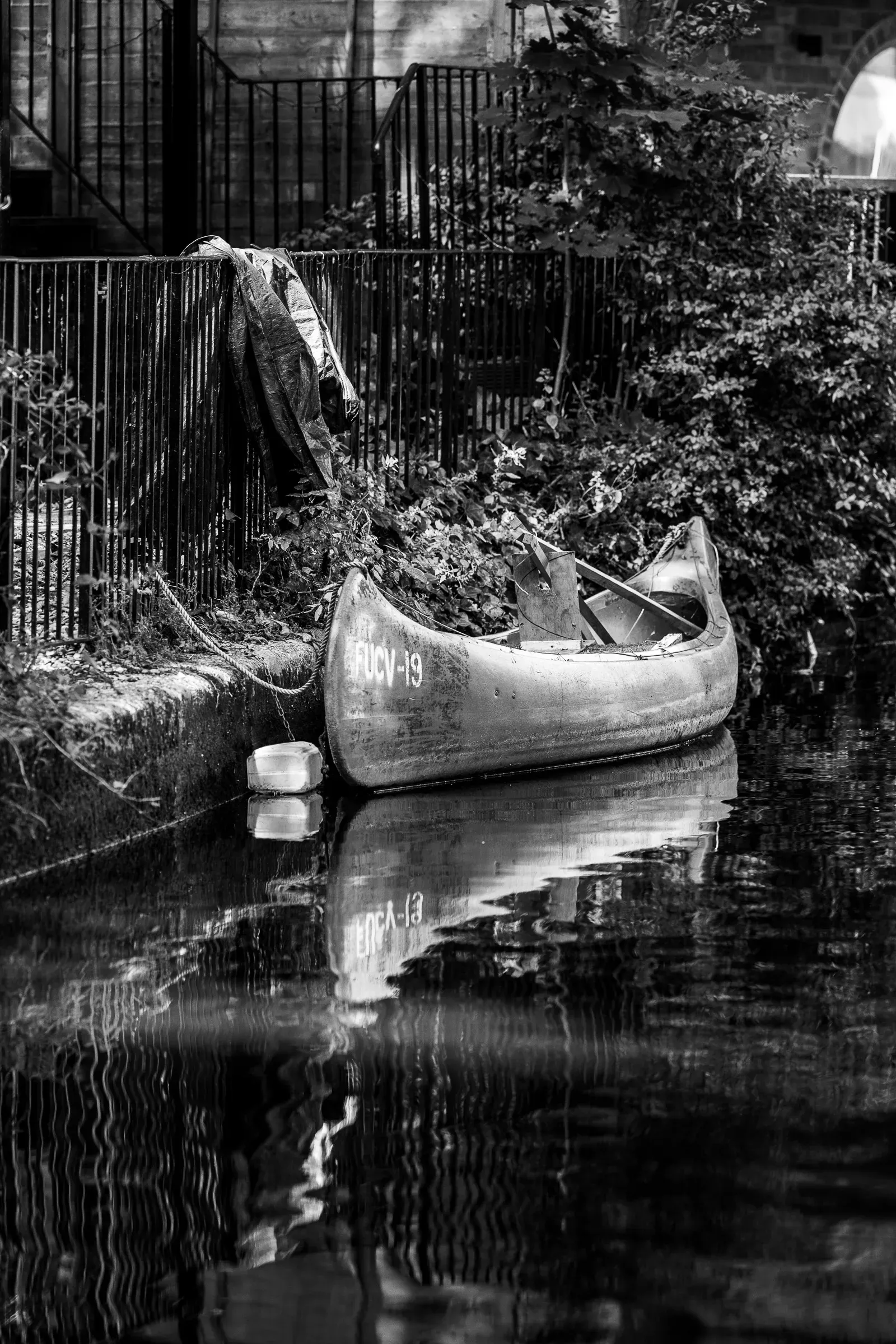 Black and white — an old canoe loaded with vegetation moored against a canal wall, iron railing above, reflected in dark water
