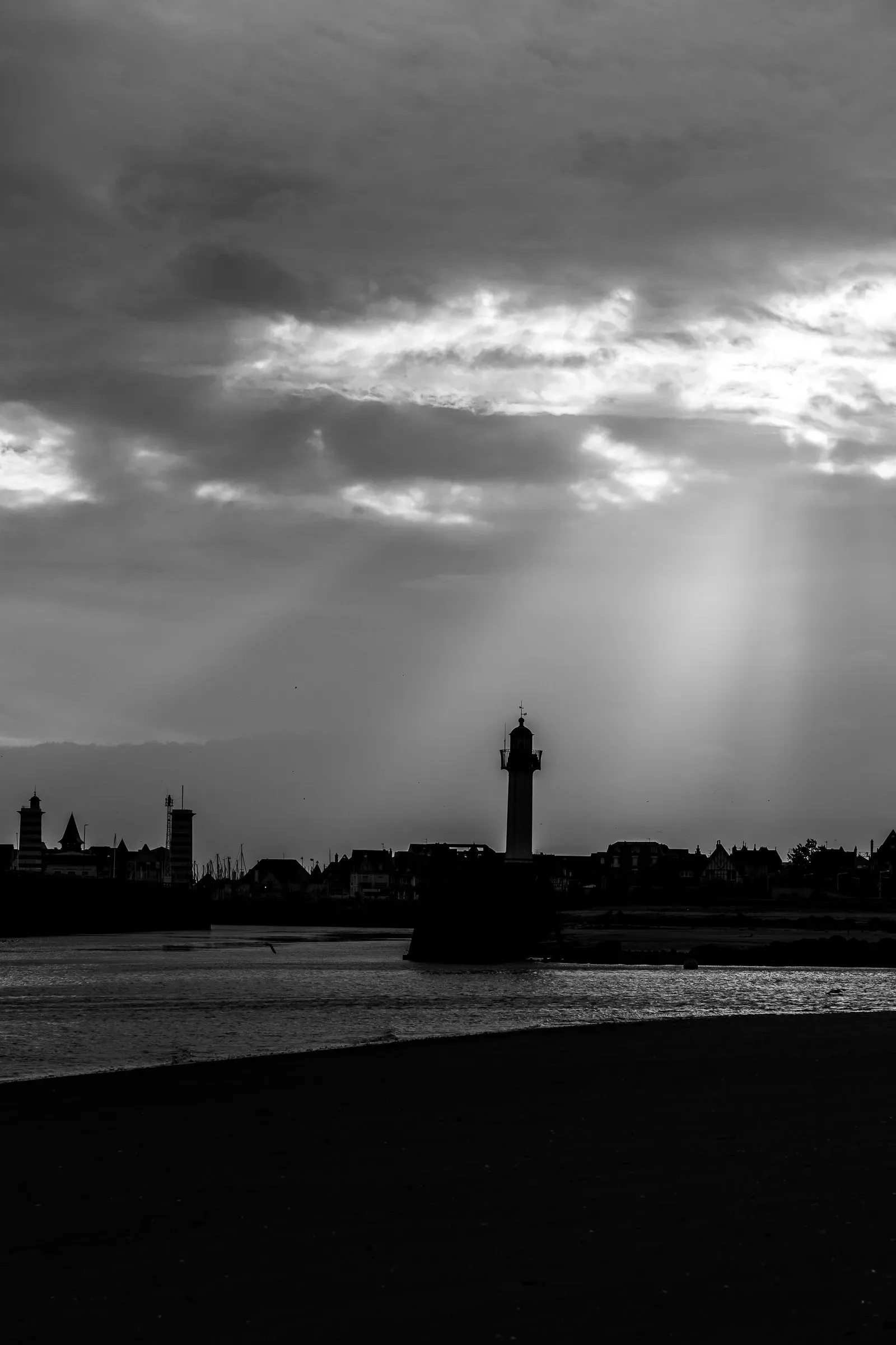 Black and white — Trouville town skyline and lighthouse silhouetted against a dramatic sky, light rays breaking through storm clouds, beach in the foreground, Trouville
