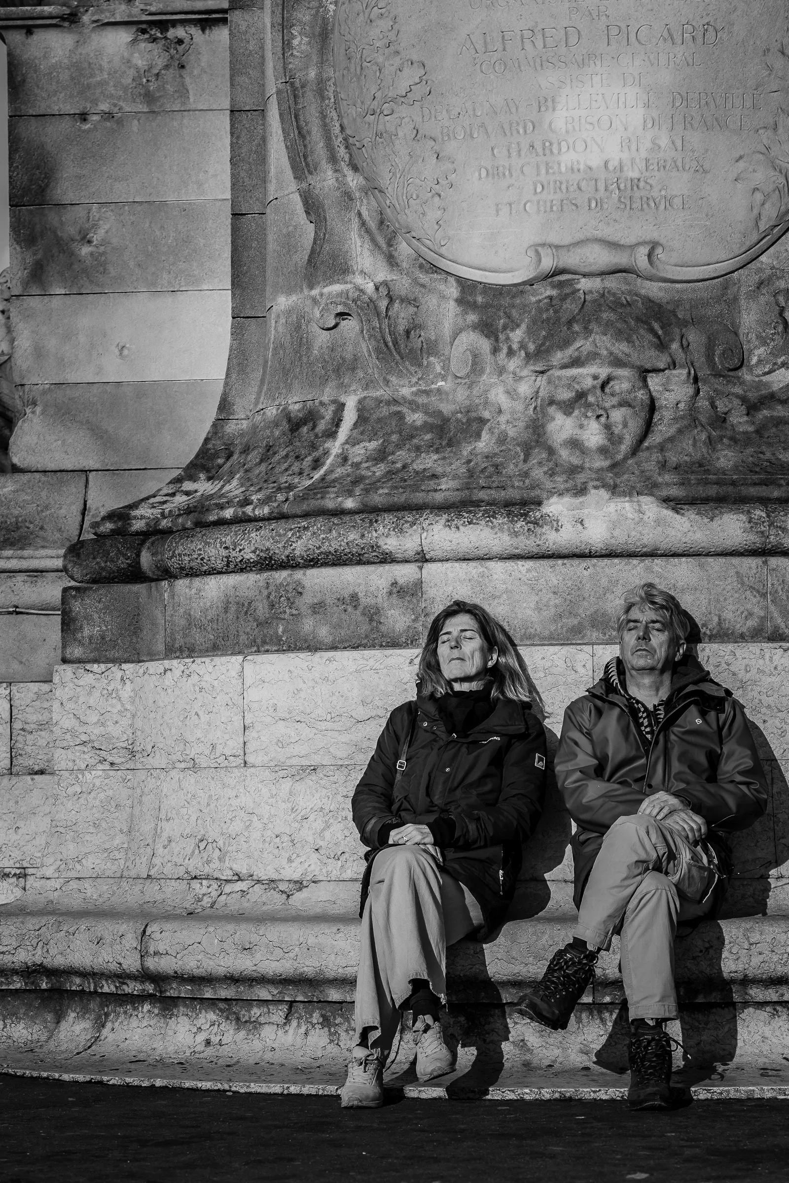 Two people sitting eyes closed in the sun at the base of a carved stone monument, an inscription naming Alfred Picard above them
