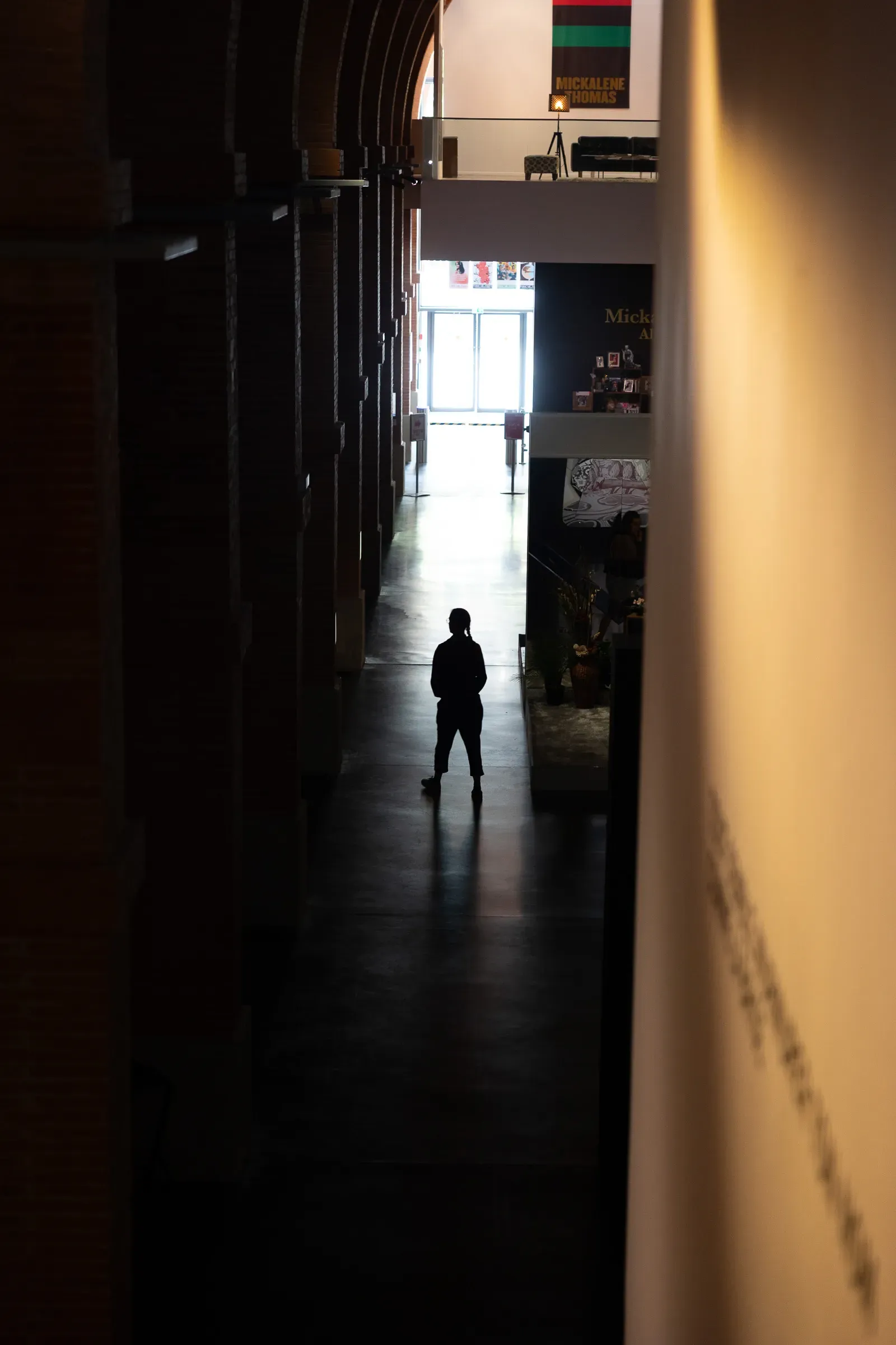 A lone silhouetted figure at the end of a long dark museum corridor, bright light flooding from the far end, a Mickalene Thomas exhibition banner visible in the background, Les Abattoirs, Toulouse