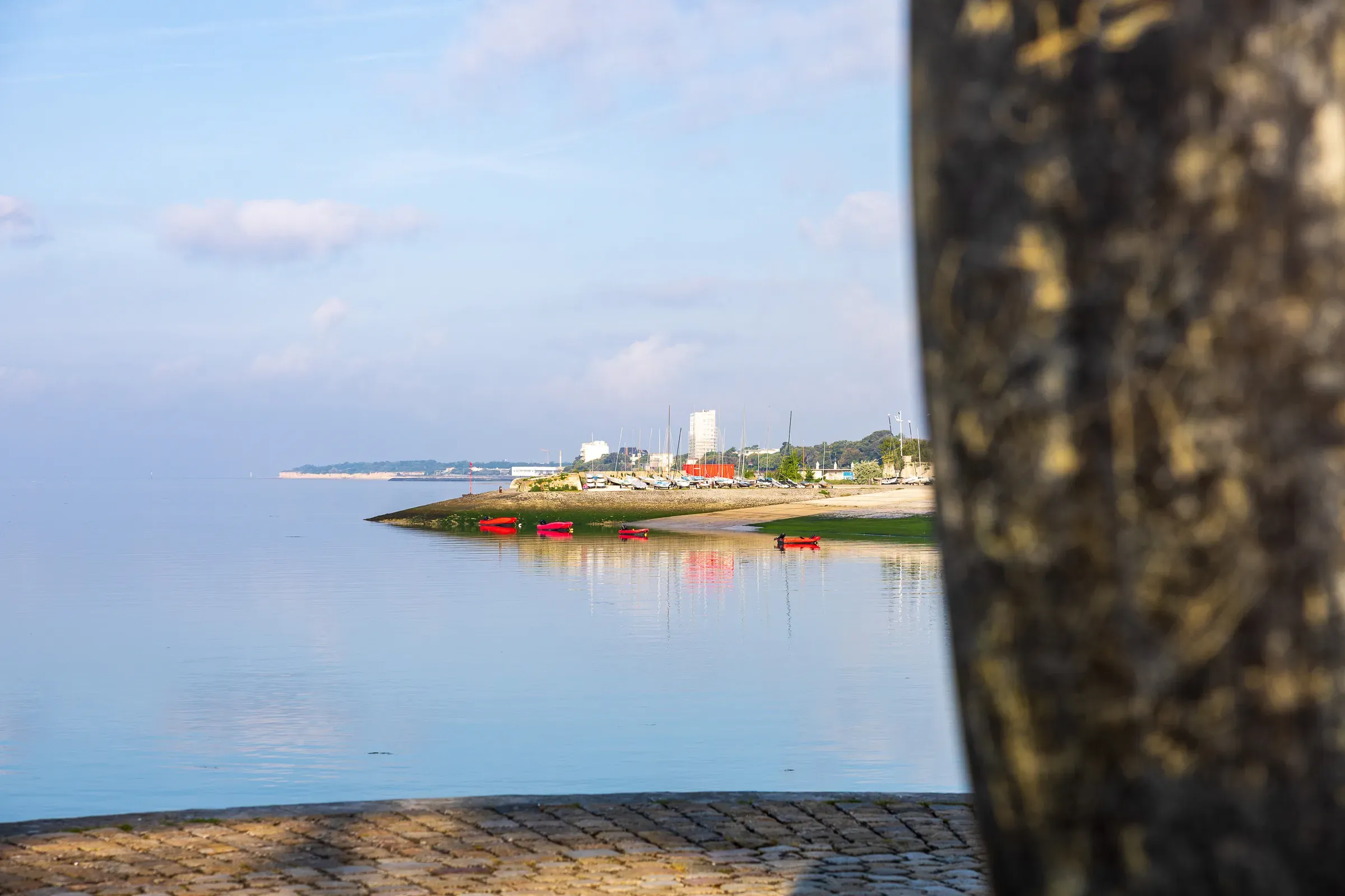 View framed between two stone walls looking onto a calm harbour at low tide, red kayaks on the shore, flat water reflecting a pale sky, distant buildings