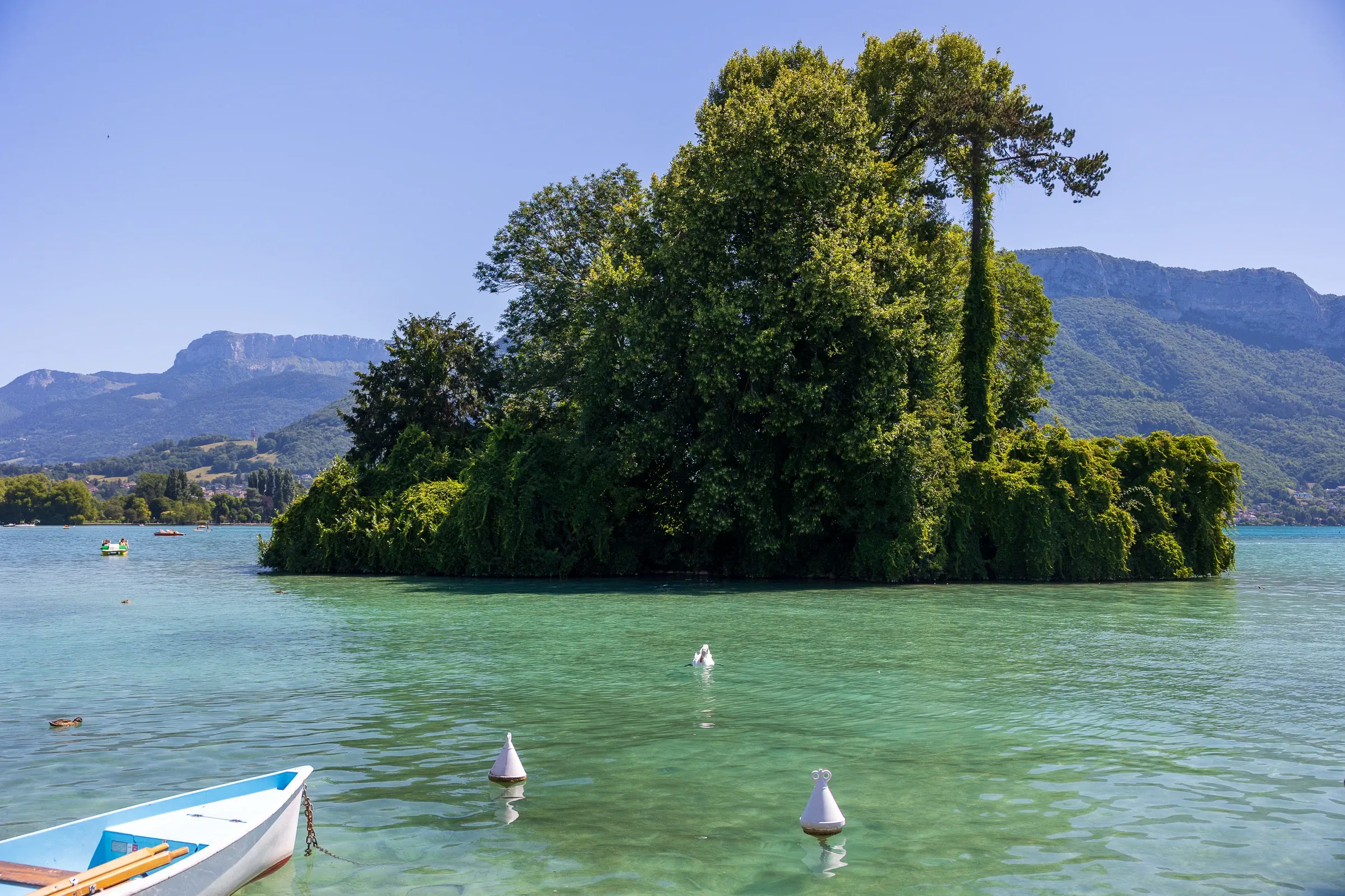 Small wooded island in Lake Annecy with forested alpine mountains and a clear blue sky