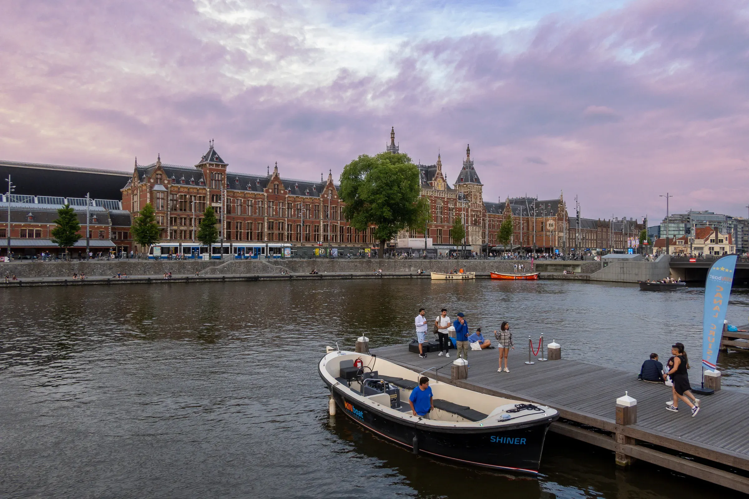 Amsterdam Centraal Station viewed across the IJ at dusk, purple-pink sky, a small boat docked in the foreground