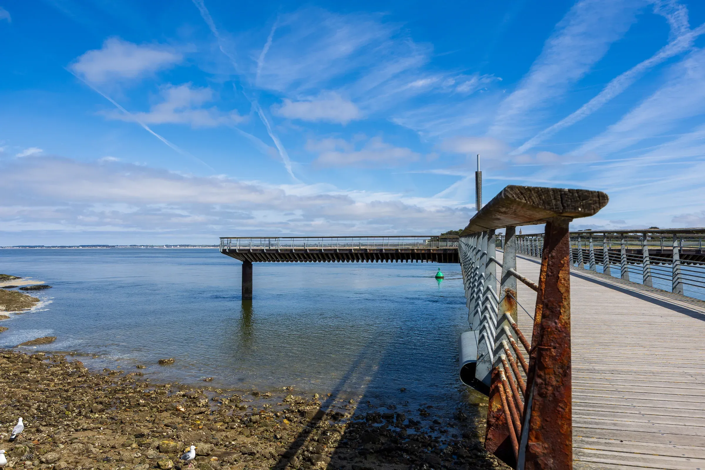 Modern rusted metal pier extending over rocky shoreline into calm blue water, contrails crossing the sky above