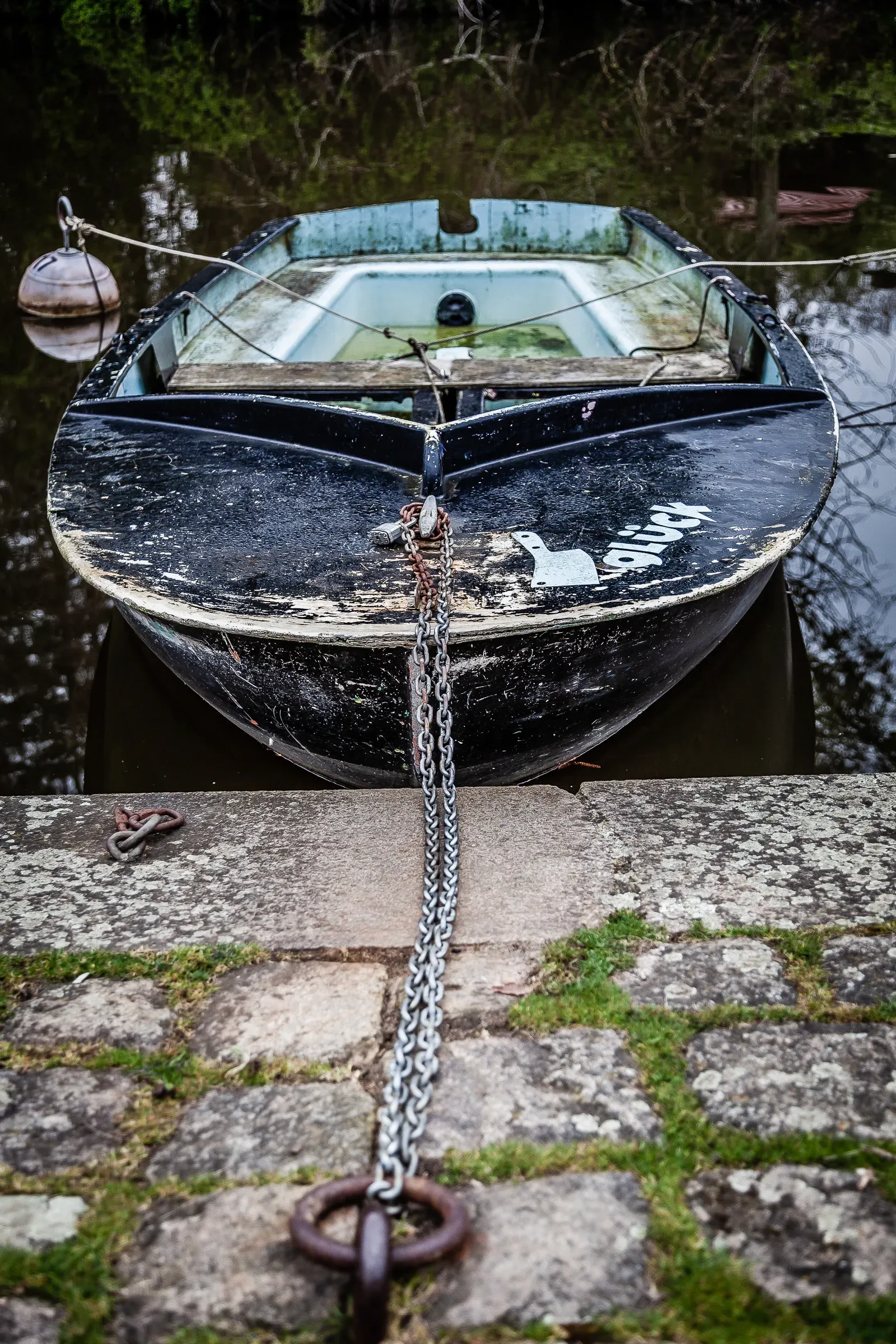 Bow of a beaten-up black boat with peeling paint, chained by a heavy link chain to a stone quay mooring ring