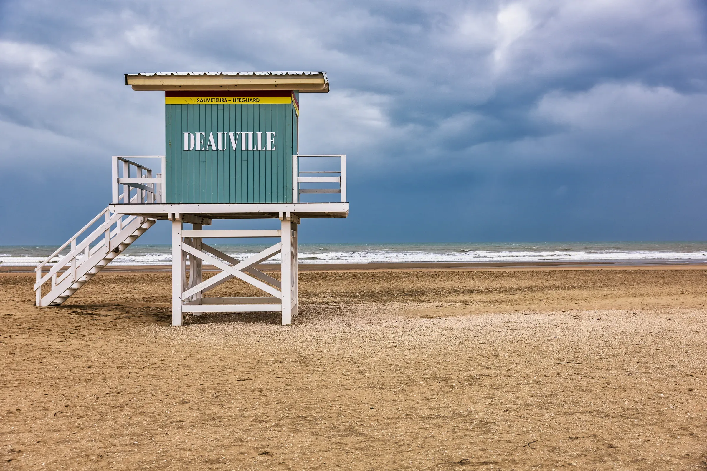 Teal lifeguard tower labeled DEAUVILLE standing alone on a wide empty winter beach under a stormy sky, Deauville