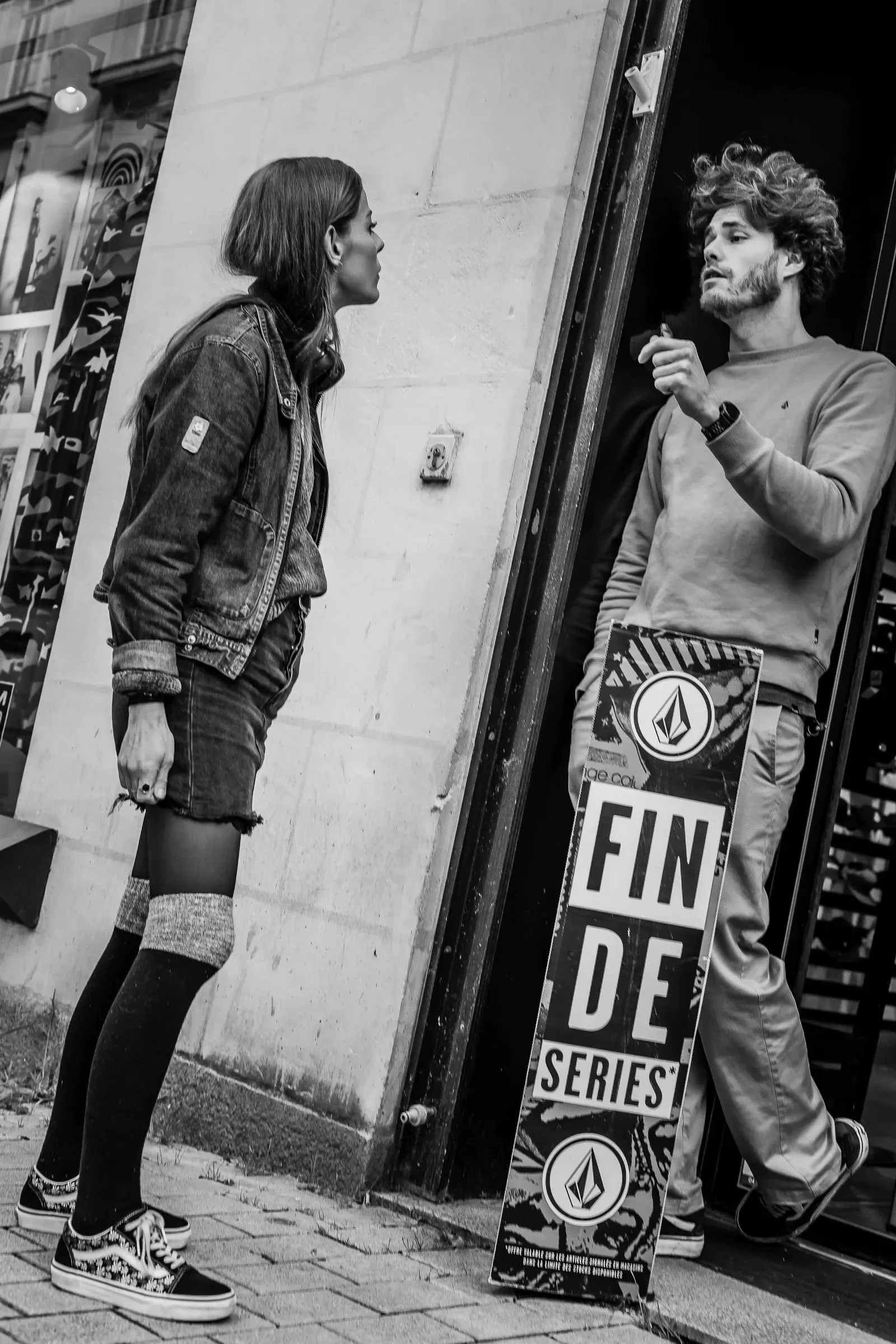 A woman glances sideways at a man smoking near a shop entrance, a 'FIN DE SERIES' sale sign propped in the doorway