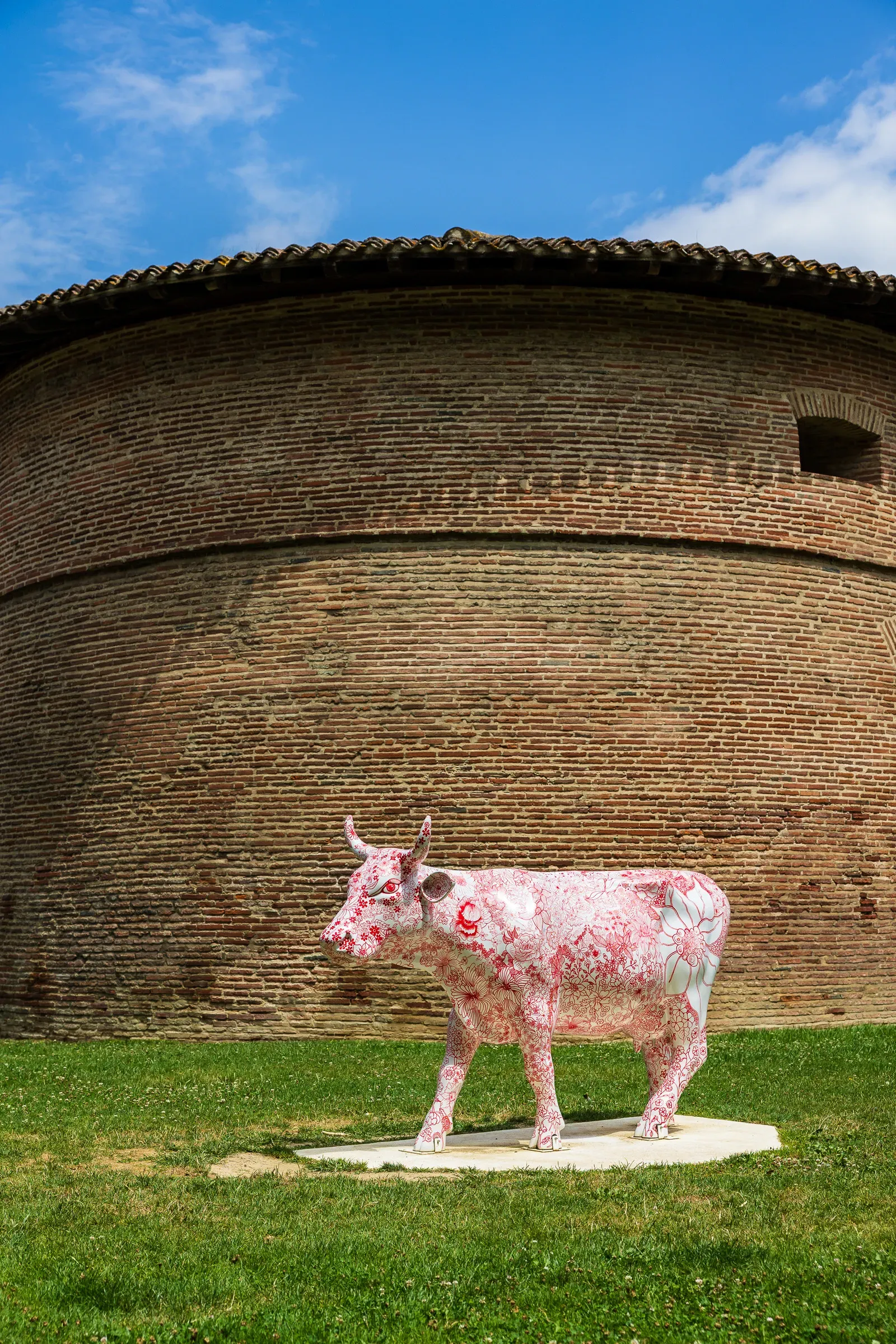 White painted cow sculpture with red floral motifs standing on grass in front of a curved red brick wall under a blue sky, Toulouse