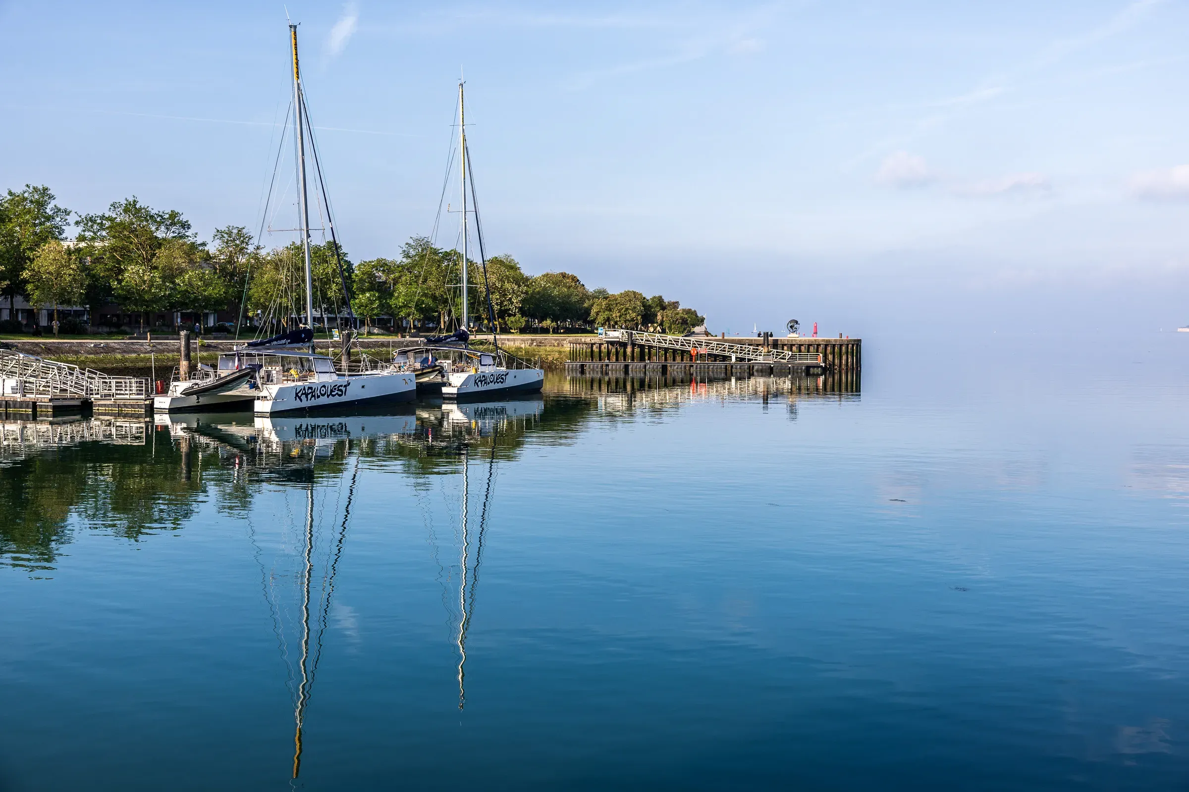 Two catamarans moored at a wooden jetty, masts perfectly mirrored in glassy morning water, tree-lined shore behind under a pale hazy sky