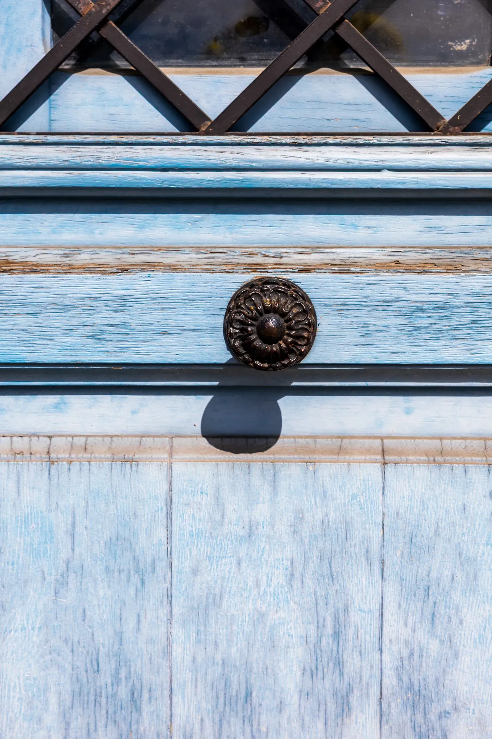 Close-up of a weathered pale blue door with an ornate iron handle and a diamond-lattice window grille above