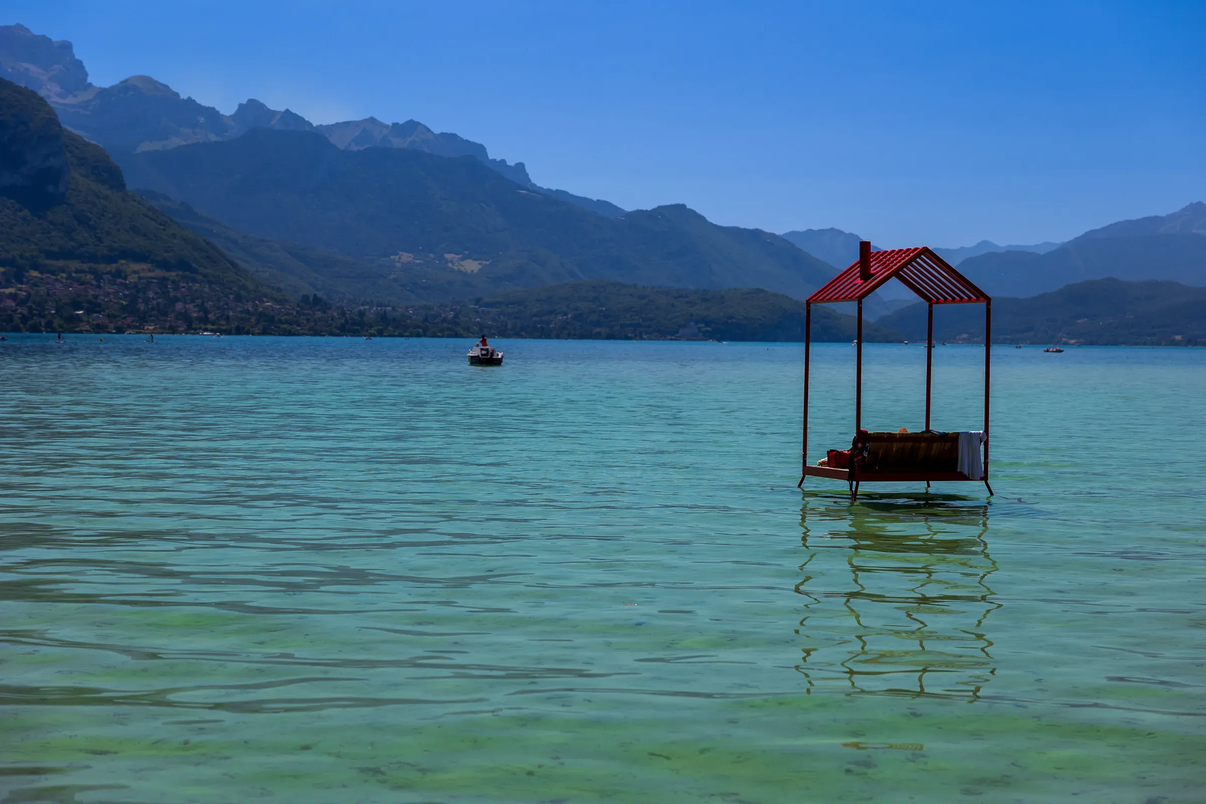 A red-roofed floating hut on the turquoise surface of Lake Annecy, mountain ridges in the background