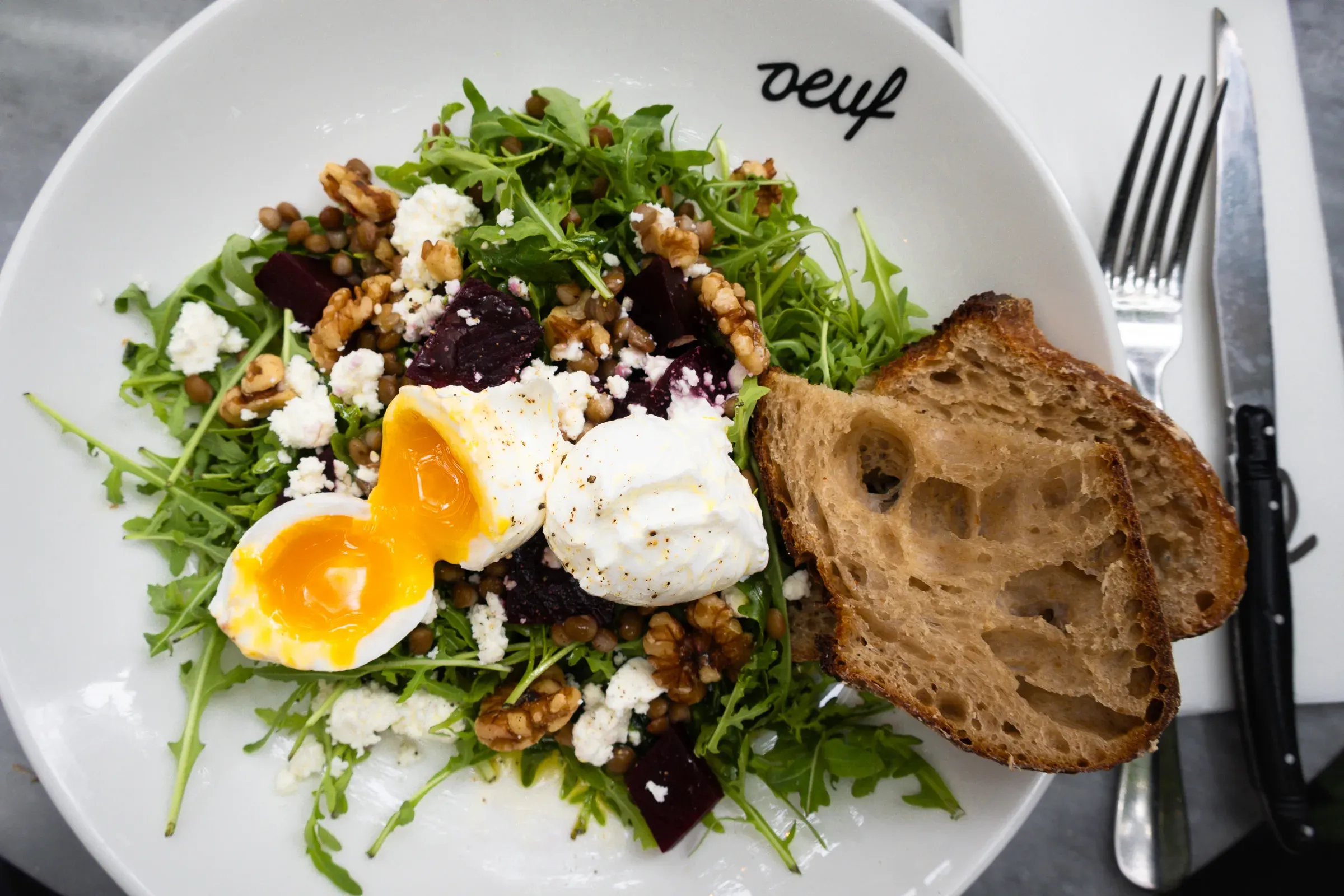 Arugula salad with soft-boiled egg, beets, goat cheese and walnuts in an oeuf-branded bowl, sourdough toast beside it