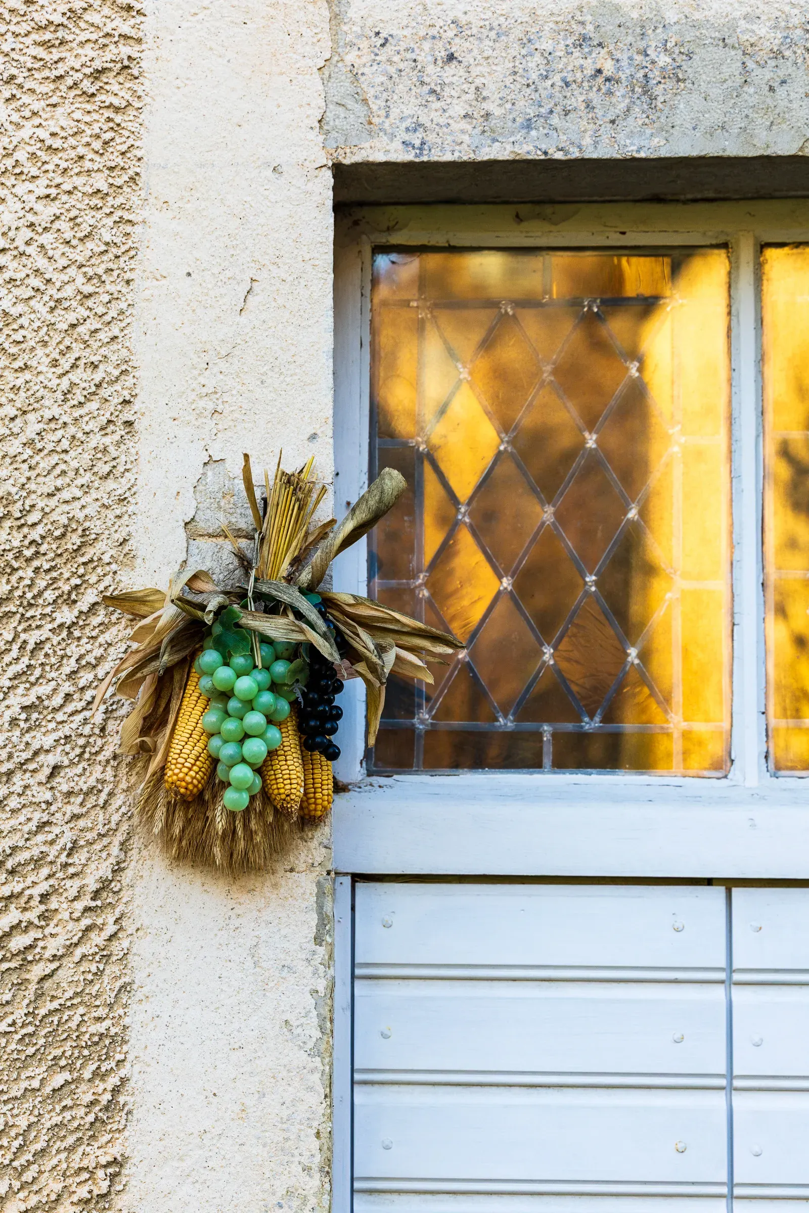 Dried corn and grape bunch hanging beside a leaded diamond-pane window glowing amber inside, Jura