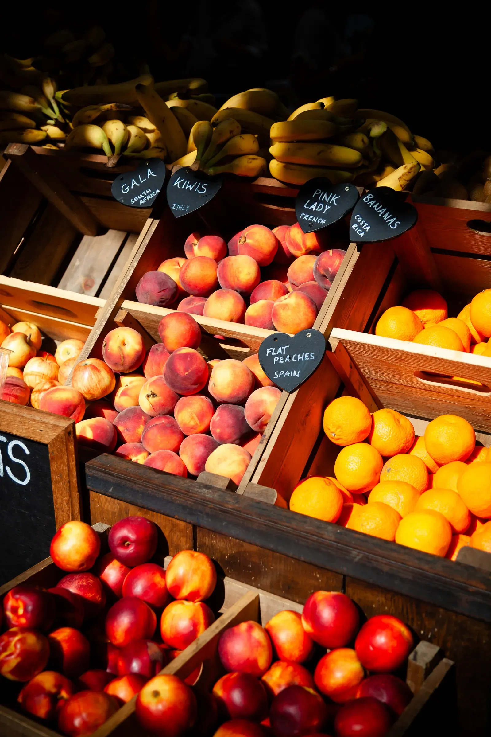 Wooden crates of flat peaches, nectarines, oranges, kiwis, and bananas at a market stall, lit with warm afternoon light and handwritten chalkboard signs