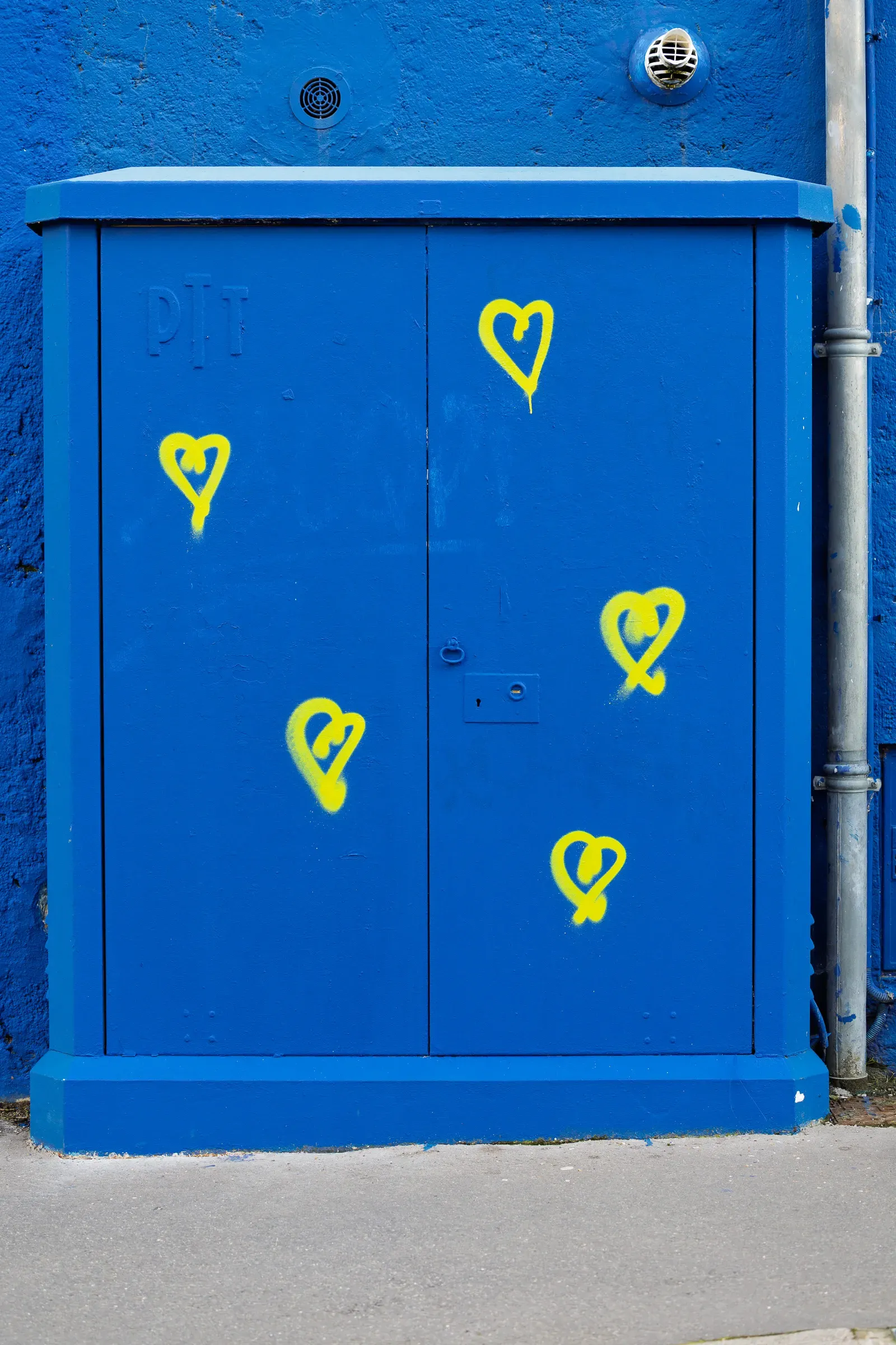 A blue utility cabinet covered in yellow spray-painted heart graffiti, set against a blue painted wall