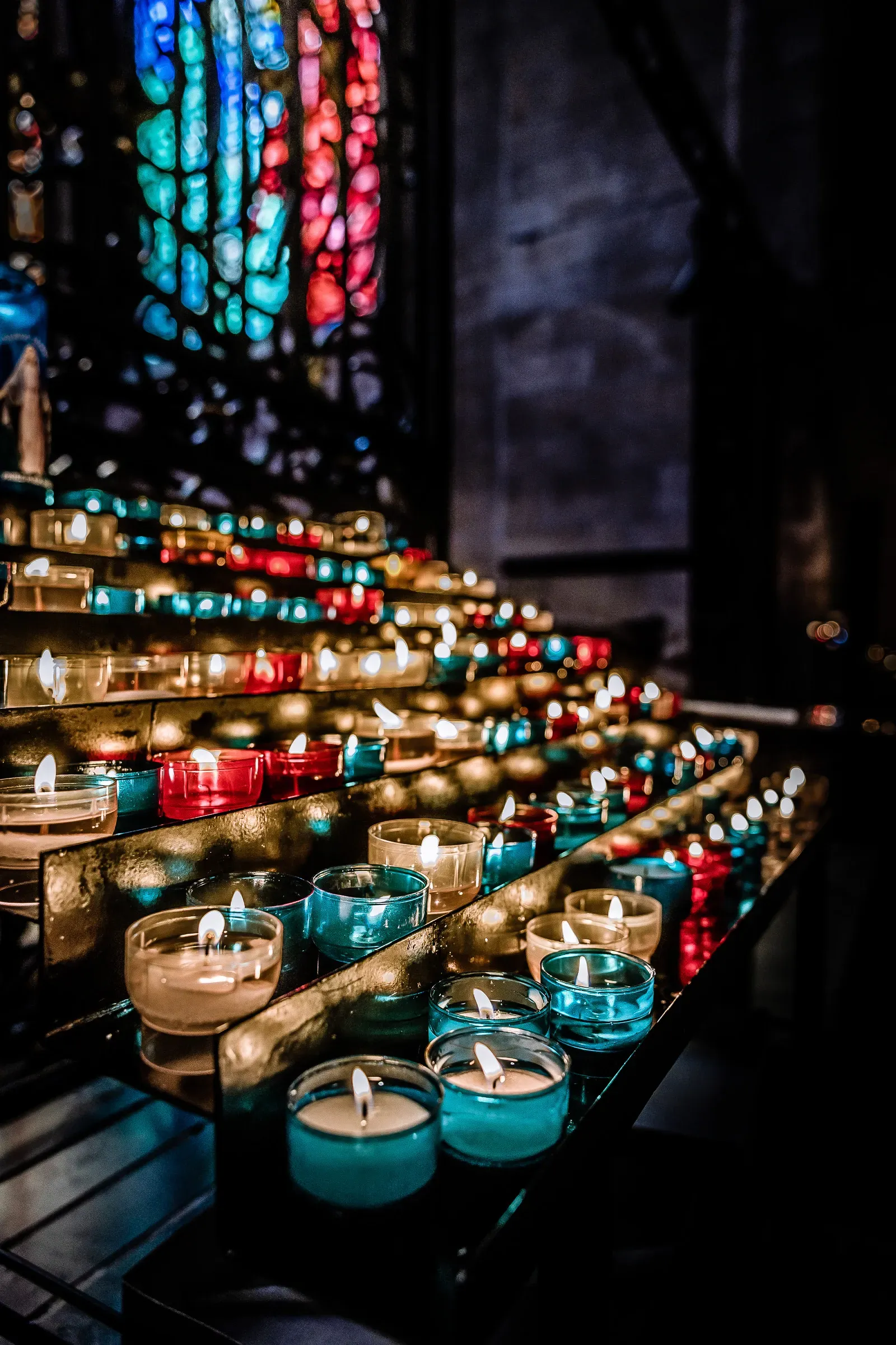 Rows of red, turquoise and gold votive candles burning inside a dark cathedral, colorful stained glass window behind