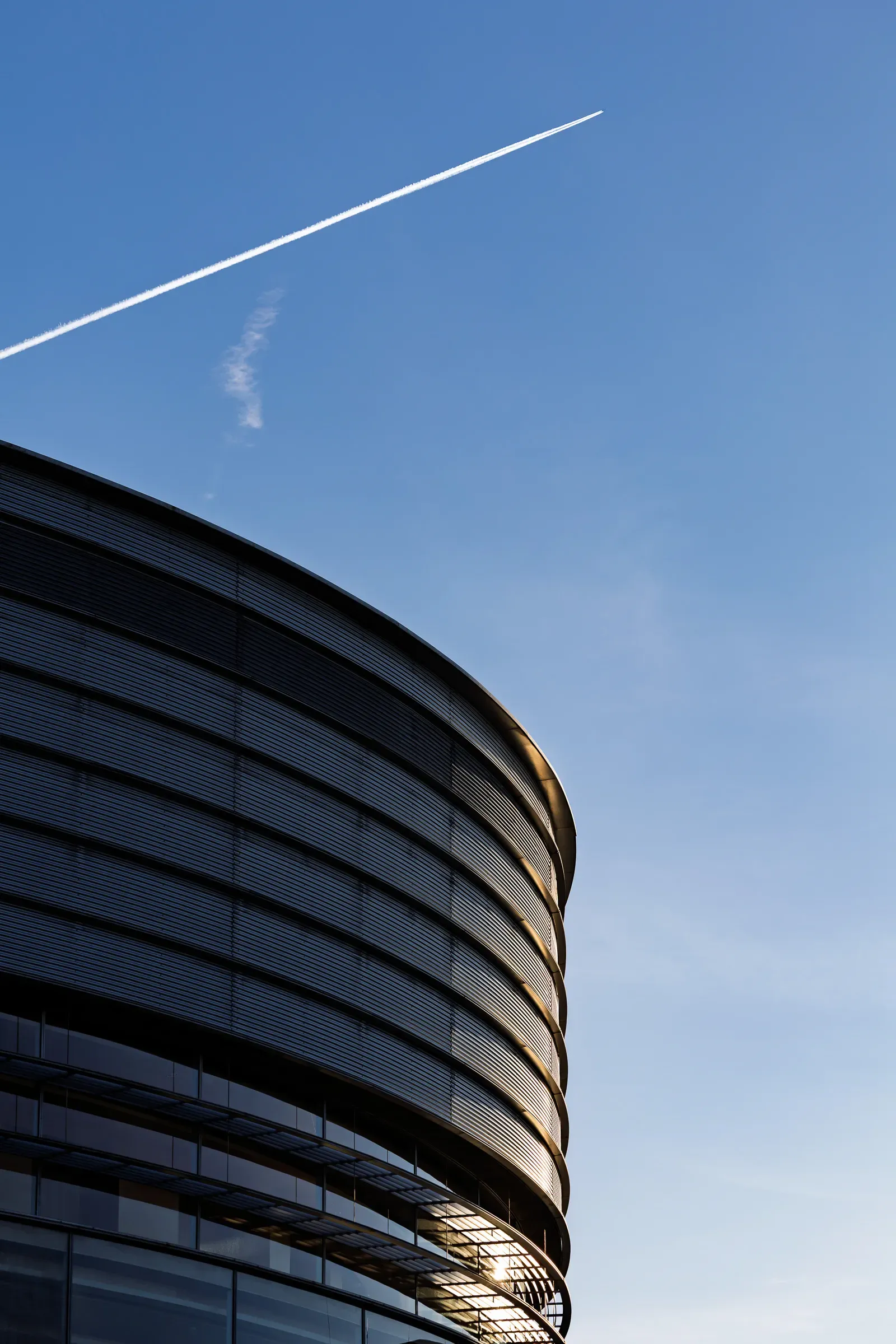 Top curve of a large circular modern building against a blue sky, a contrail crossing diagonally above