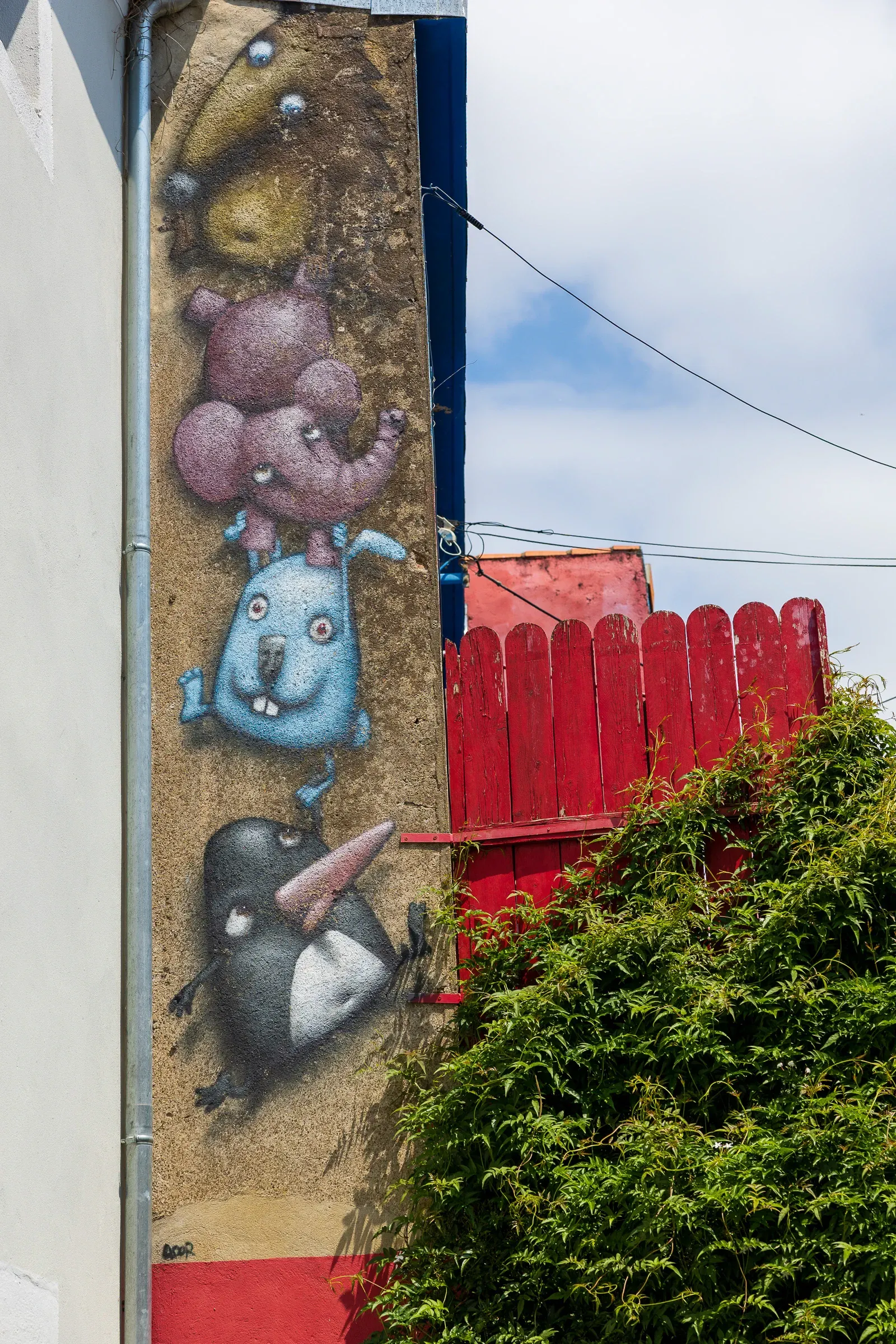 Tall street-art mural of stacked cartoon monster creatures on a house gable, red fence and greenery alongside