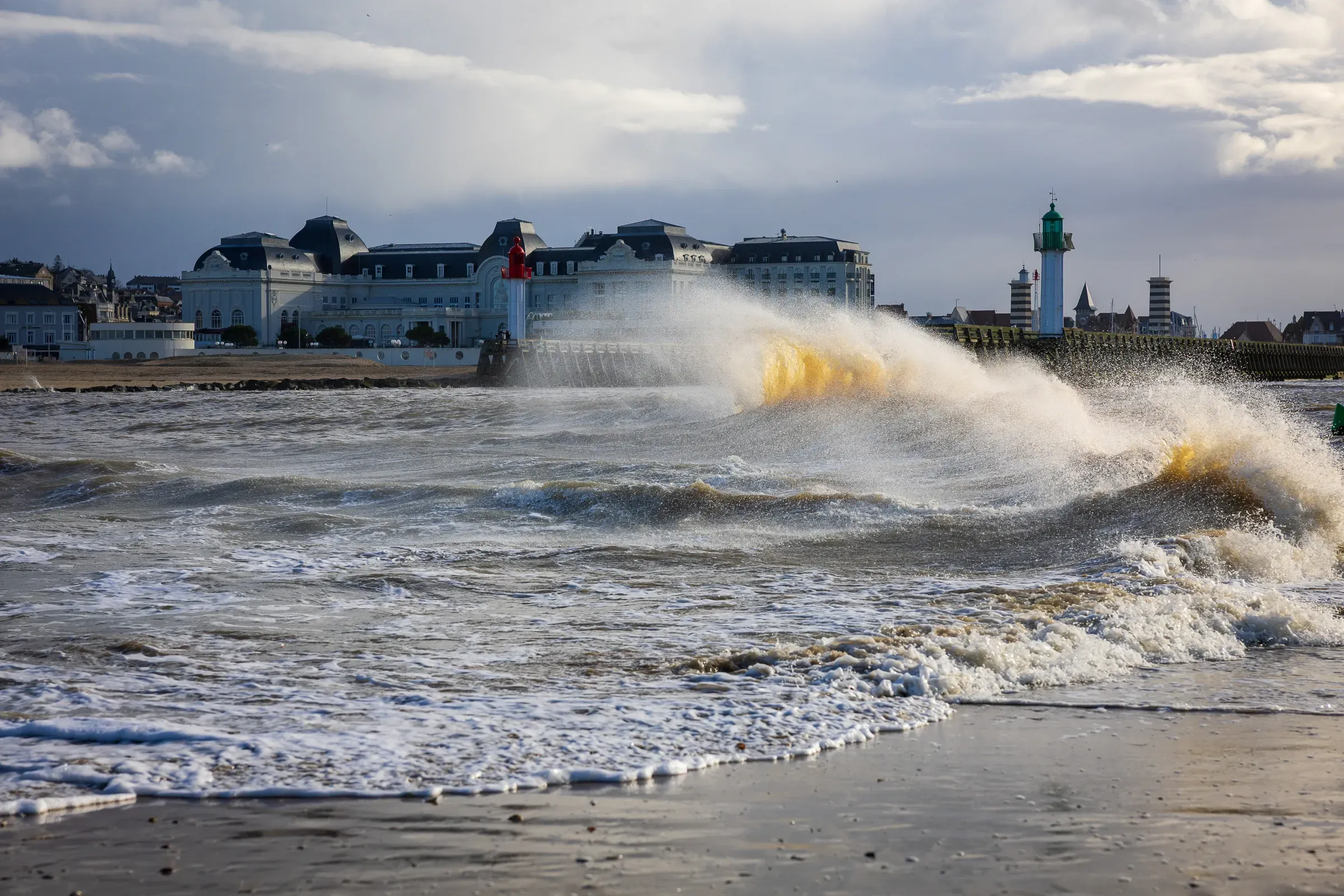 Wave crashing against the jetty wall with yellow sea spray, Trouville Casino and town buildings behind, green lighthouse to the right, Trouville