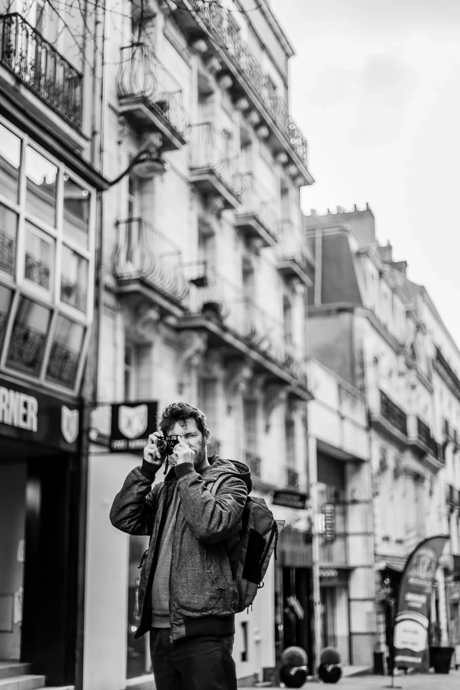 A man with a backpack raises his camera to photograph the street, buildings blurred behind him