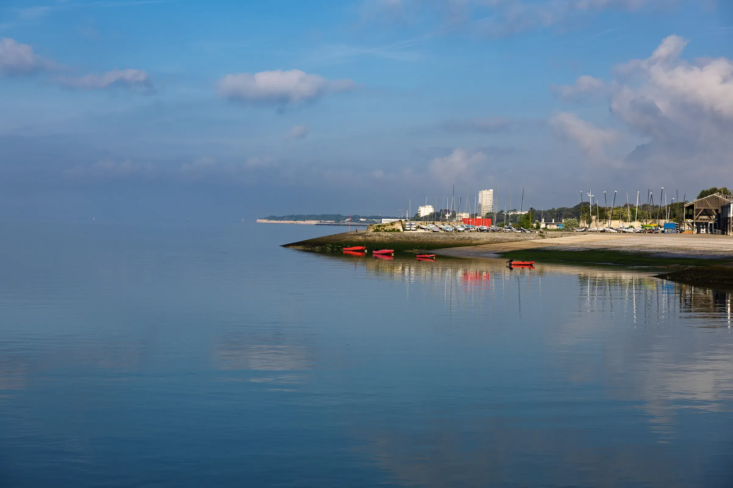 Red kayaks on a sandy spit at low tide, perfectly calm water reflecting scattered clouds, marina and city buildings stretching across the background