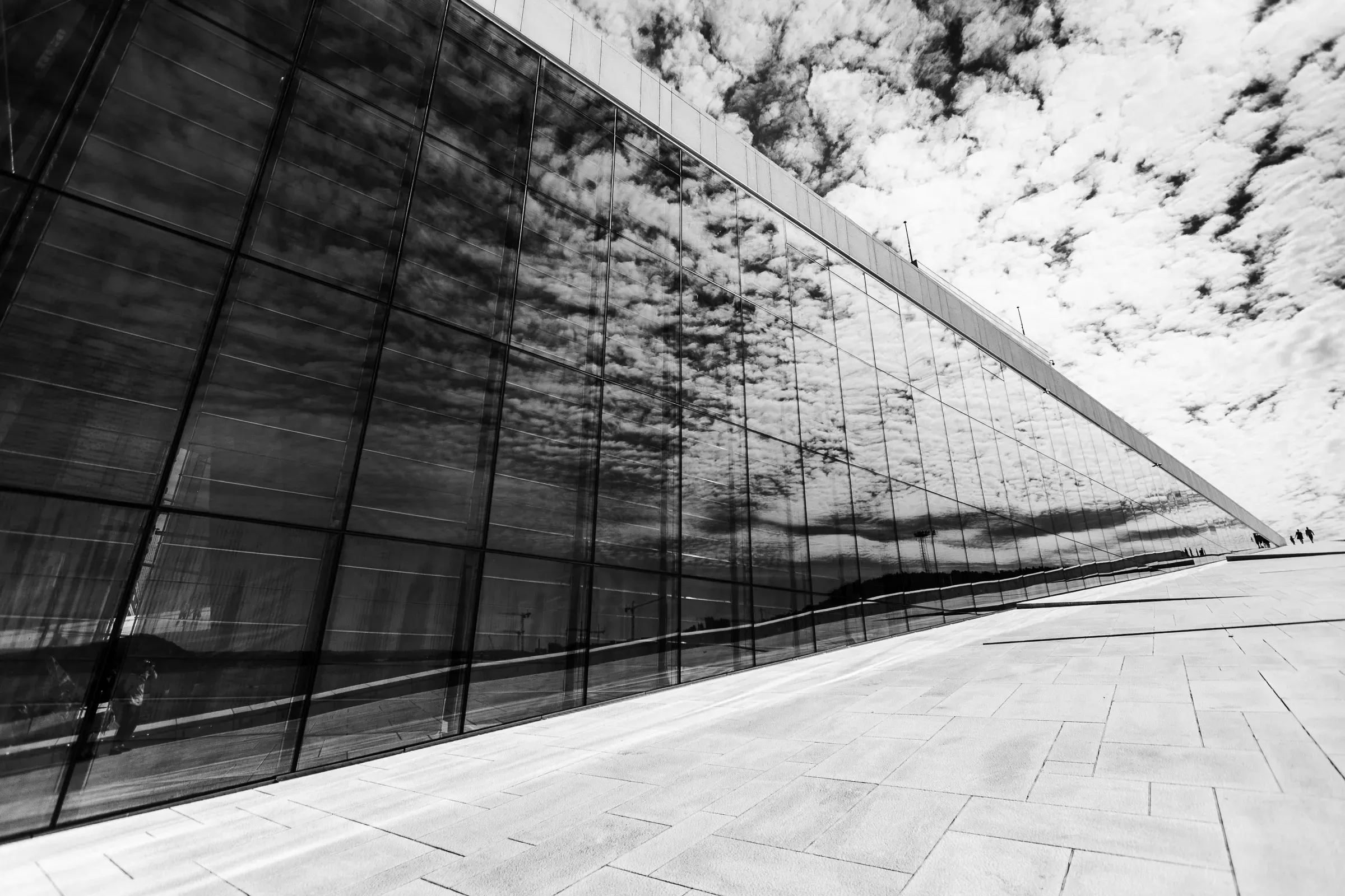 Oslo Opera House glass facade stretching diagonally across the frame, clouds and sky reflected along its full length, tiny figures at the far edge