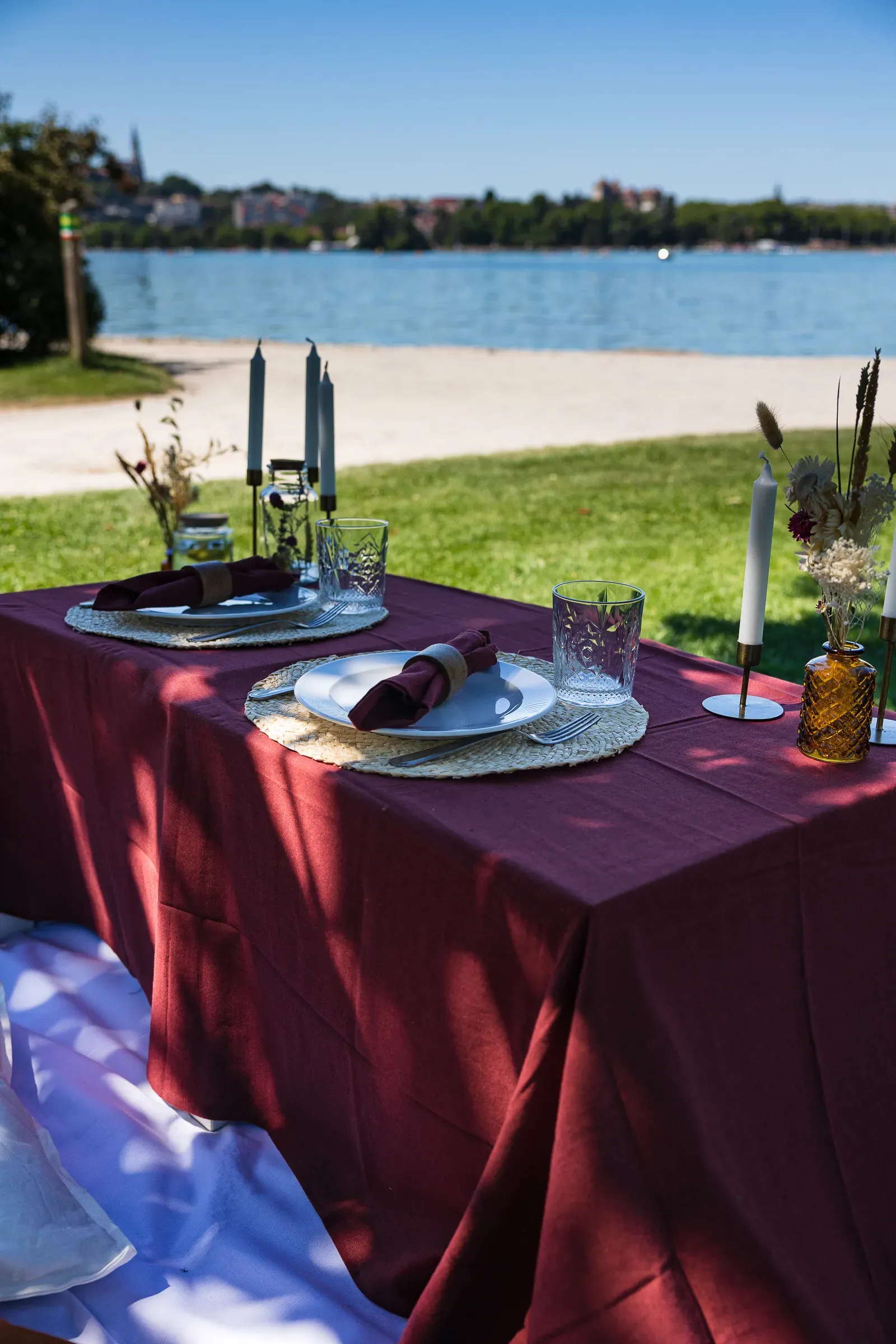 A table set with burgundy linen and candles on a lakeside lawn, Lake Annecy behind in bright sun