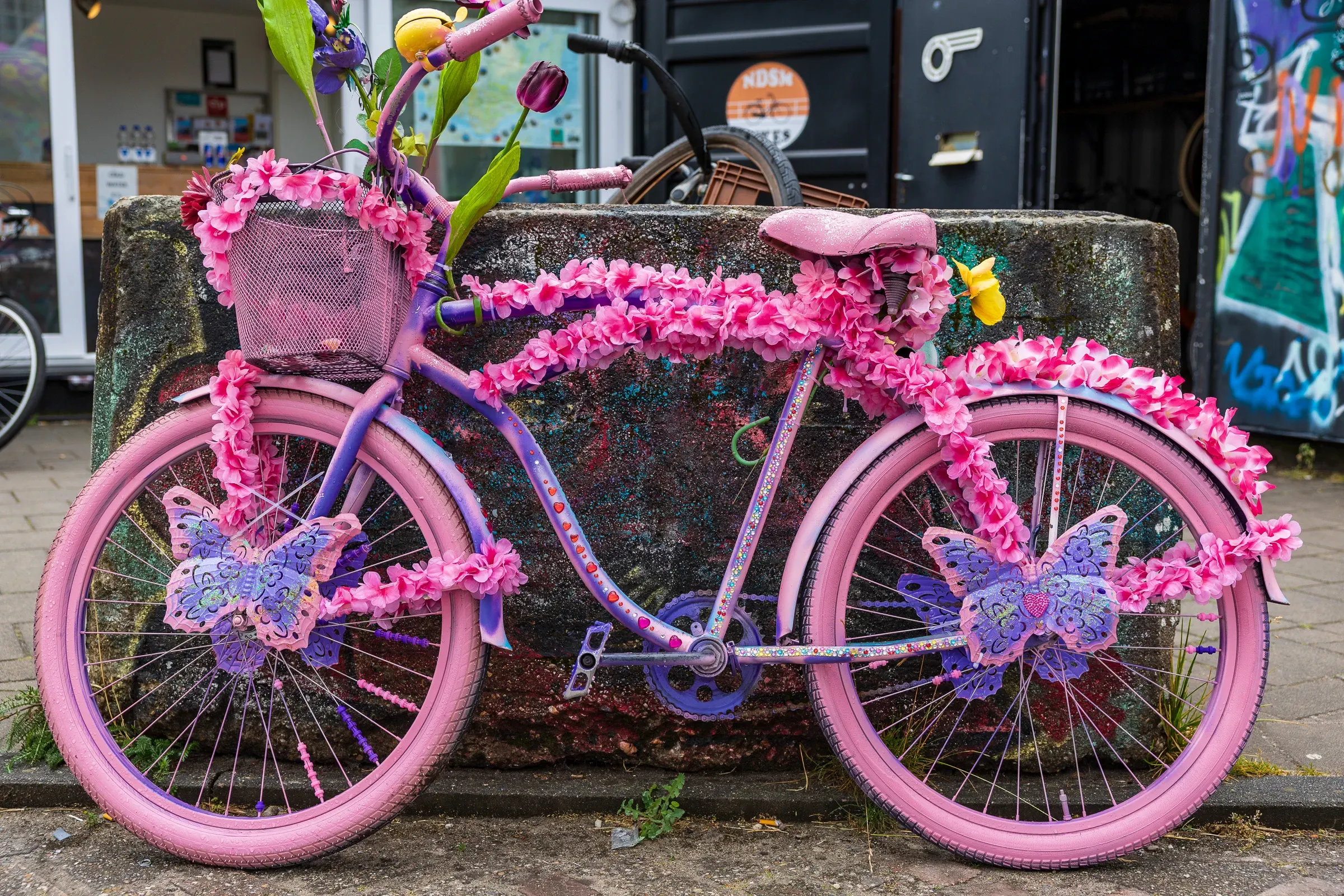 An all-pink bicycle covered in pink fabric flowers and butterfly ornaments, leaning against a graffiti-covered concrete block