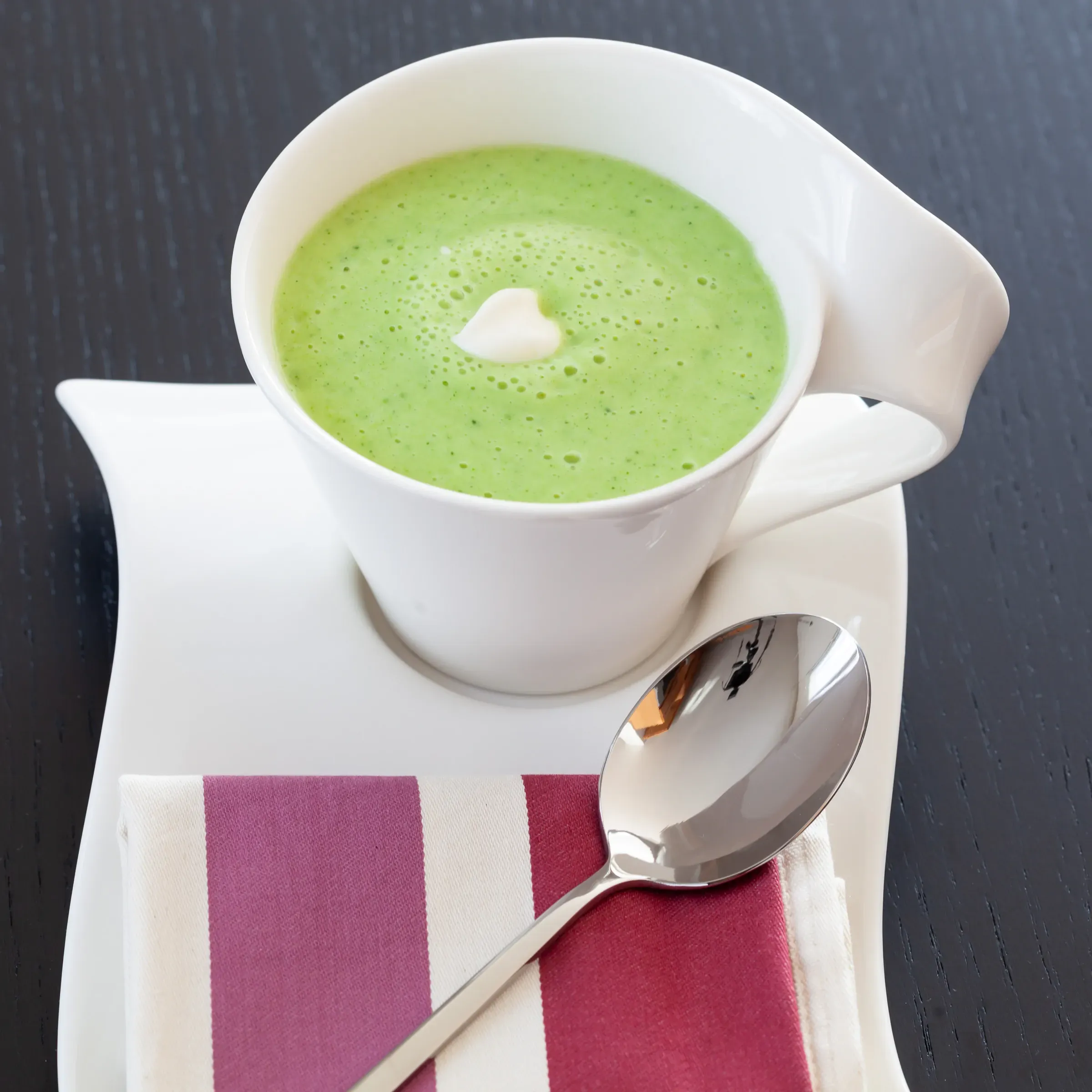 Pea velouté in a white cup, single drop of cream at the centre, striped cloth and silver spoon on a dark surface
