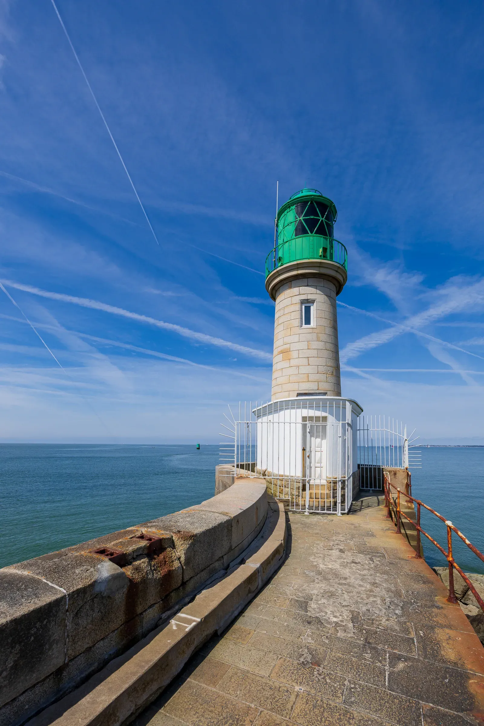 Green-capped granite lighthouse at the end of a stone jetty, open sea and blue sky behind