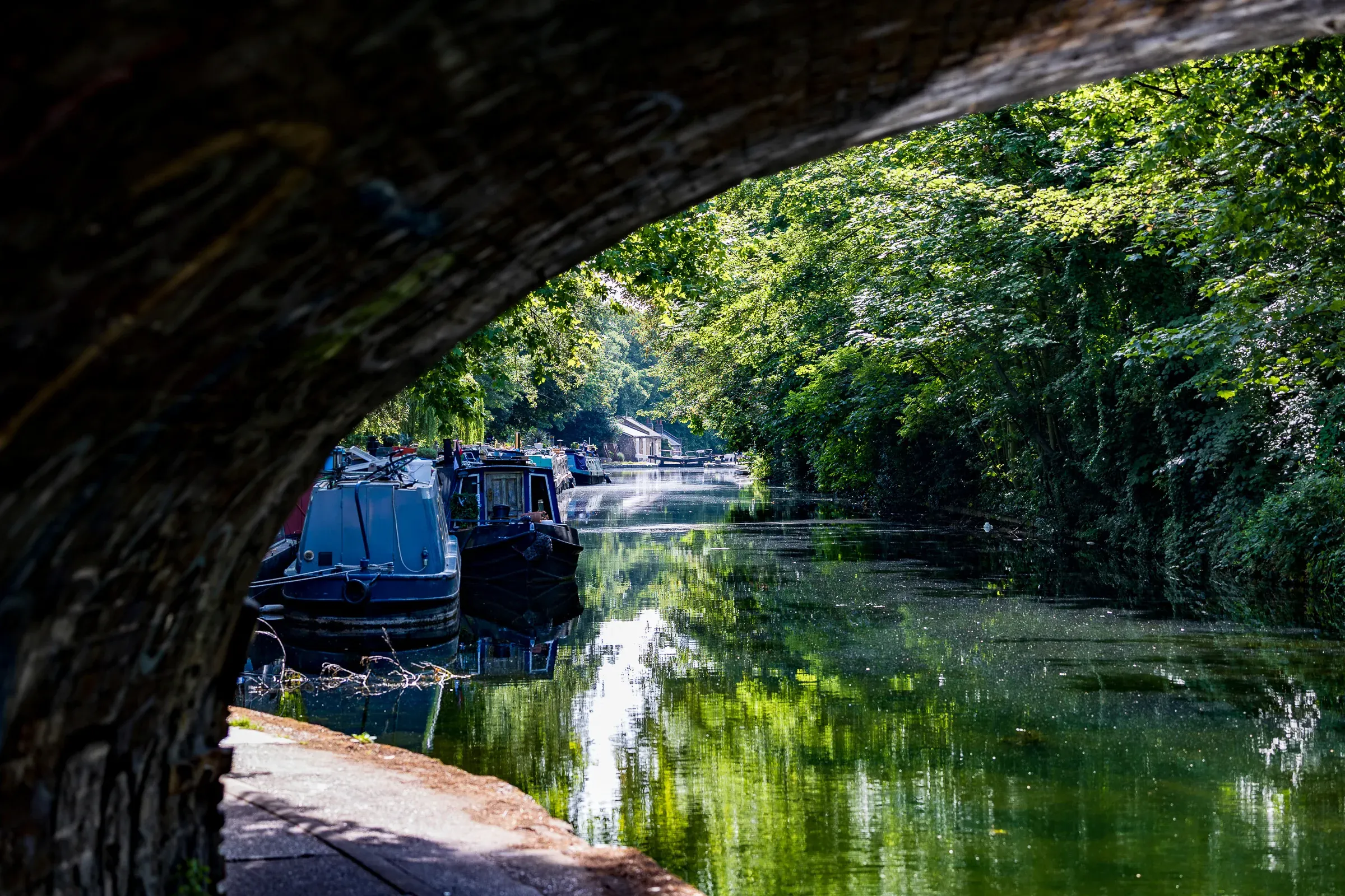 View through a brick arch onto the Regent's Canal, narrowboats moored to the left, green canopy overhead, light at the far end