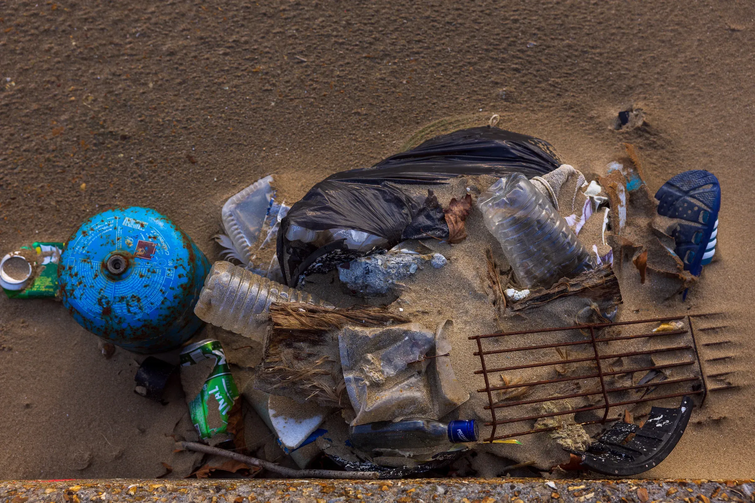 Overhead close-up of washed-up debris — plastic bottles, a blue gas canister, a black bin bag and a rusted metal grate on wet sand, Trouville
